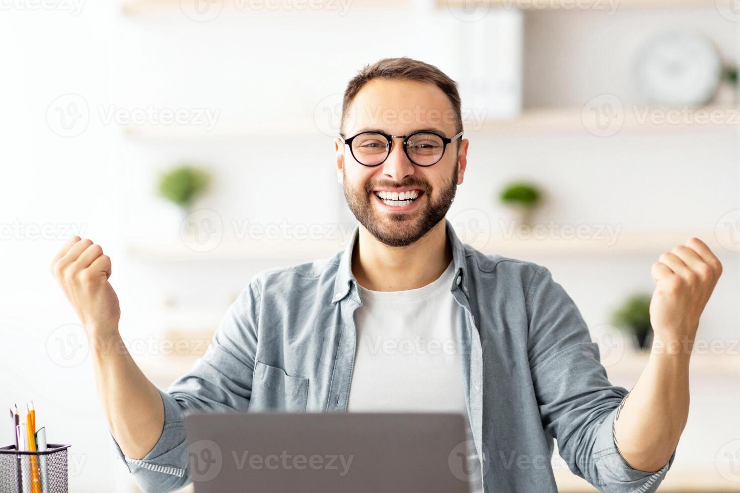 Young guy making YES gesture in front of laptop at home office. Millennial man reaching success, working with portable pc, making profitable deal, signing online contract, celebrating achievement photo