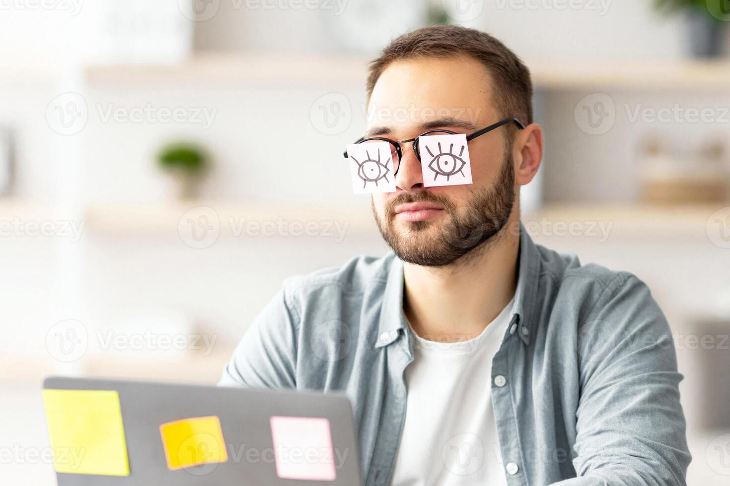 Tired millennial guy having stickers with drawings of open eyes on his glasses, sleeping in front of laptop at home office. Exhausted young man overworking, falling asleep a workplace photo