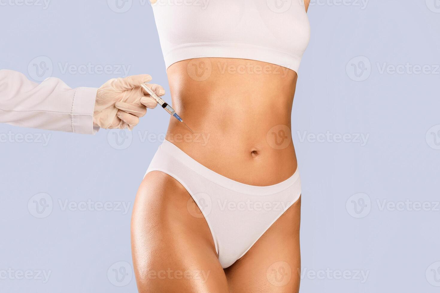 Beauty Procedure For Perfect Body. Slim woman having lipolysis treatment at beauty salon clinic. Plastic surgeon in gloves making injection at belly area for young lady, blue studio background photo