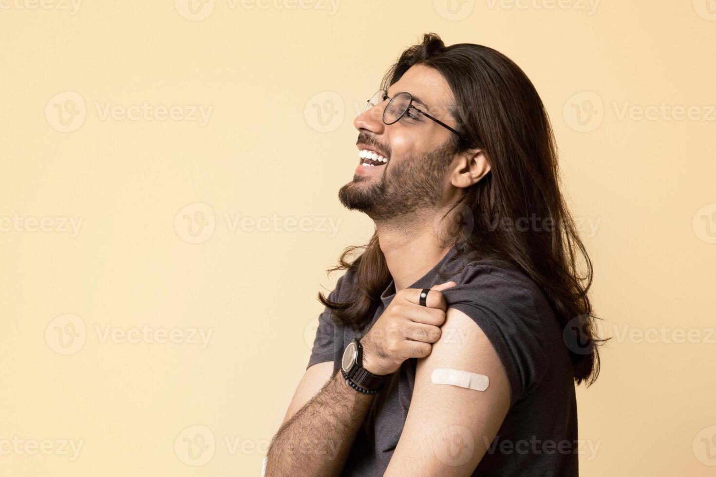 Positive middle-eastern long-haired young man in casual sitting on chair over studio background, got vaccinated against coronavirus, showing plaster, looking at copy space and smiling, panorama photo