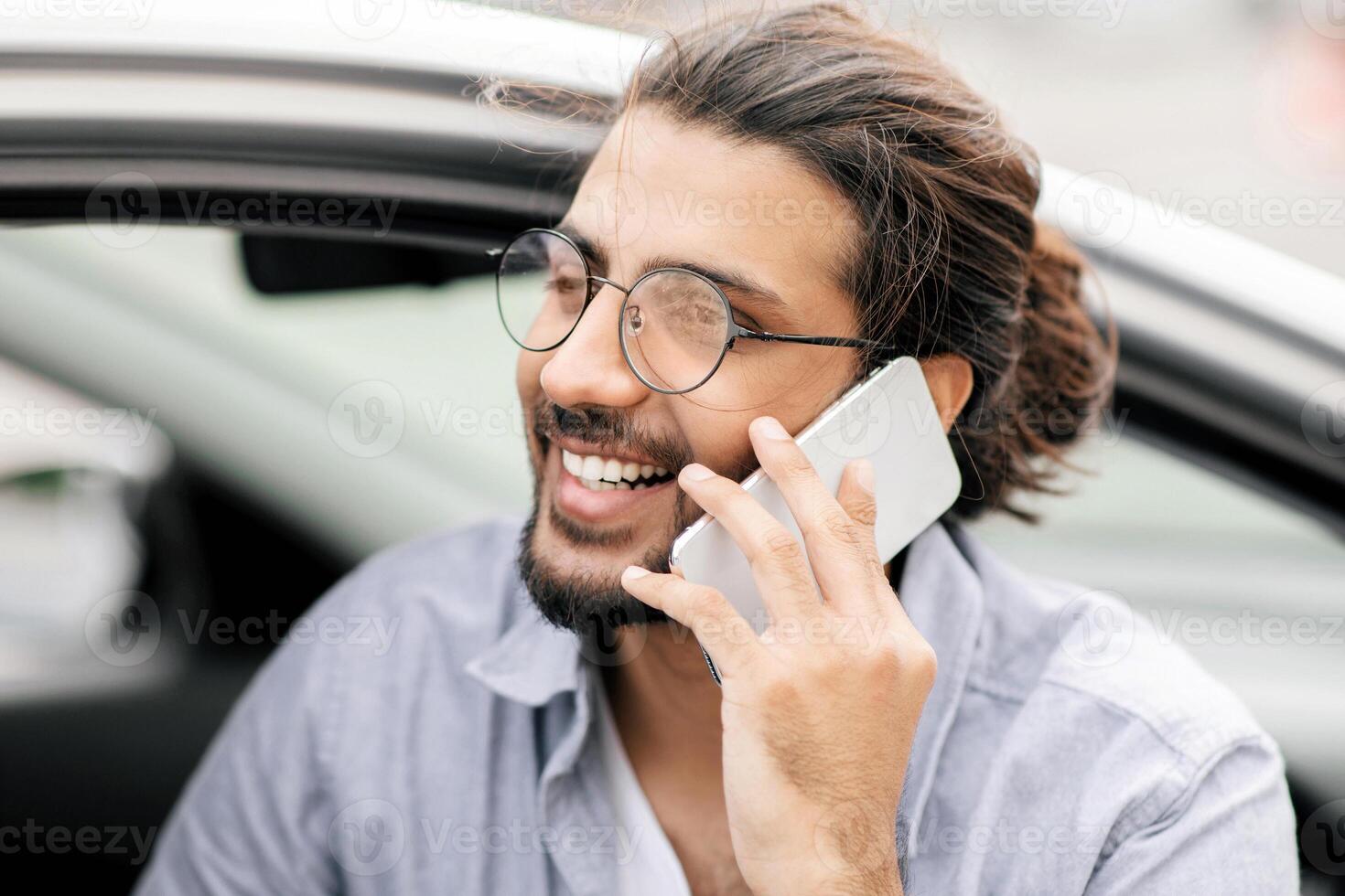 Closeup portrait of young cheerful middle-eastern man driver having phone call while driving car, side view, copy space. Happy arab guy enjoying summer trip, sitting in auto, talking on smartphone photo