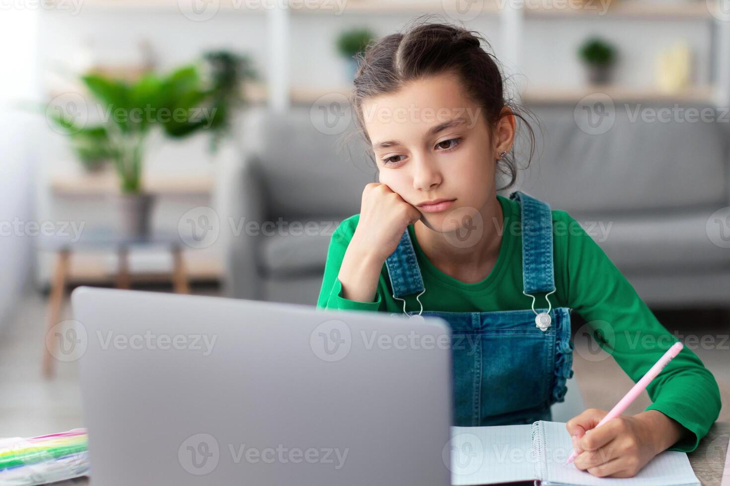 Too Tired For School. Portrait of bored upset student girl sitting at desk, doing homework, writing in her notebook and looking at laptop screen, studying online, selective focus. Home Based Learning photo