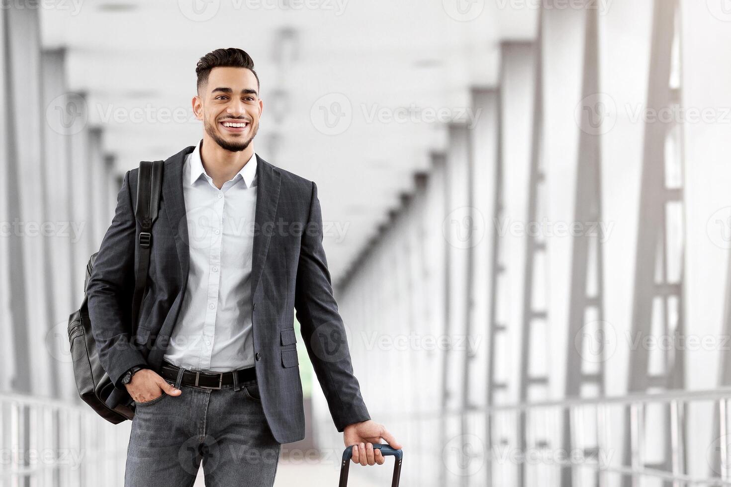 Portrait of young middle eastern man walking in airport terminal with luggage, smiling arab guy carrying suitcase while going to boarding gate, enjoying air travels and business trips, copy space photo