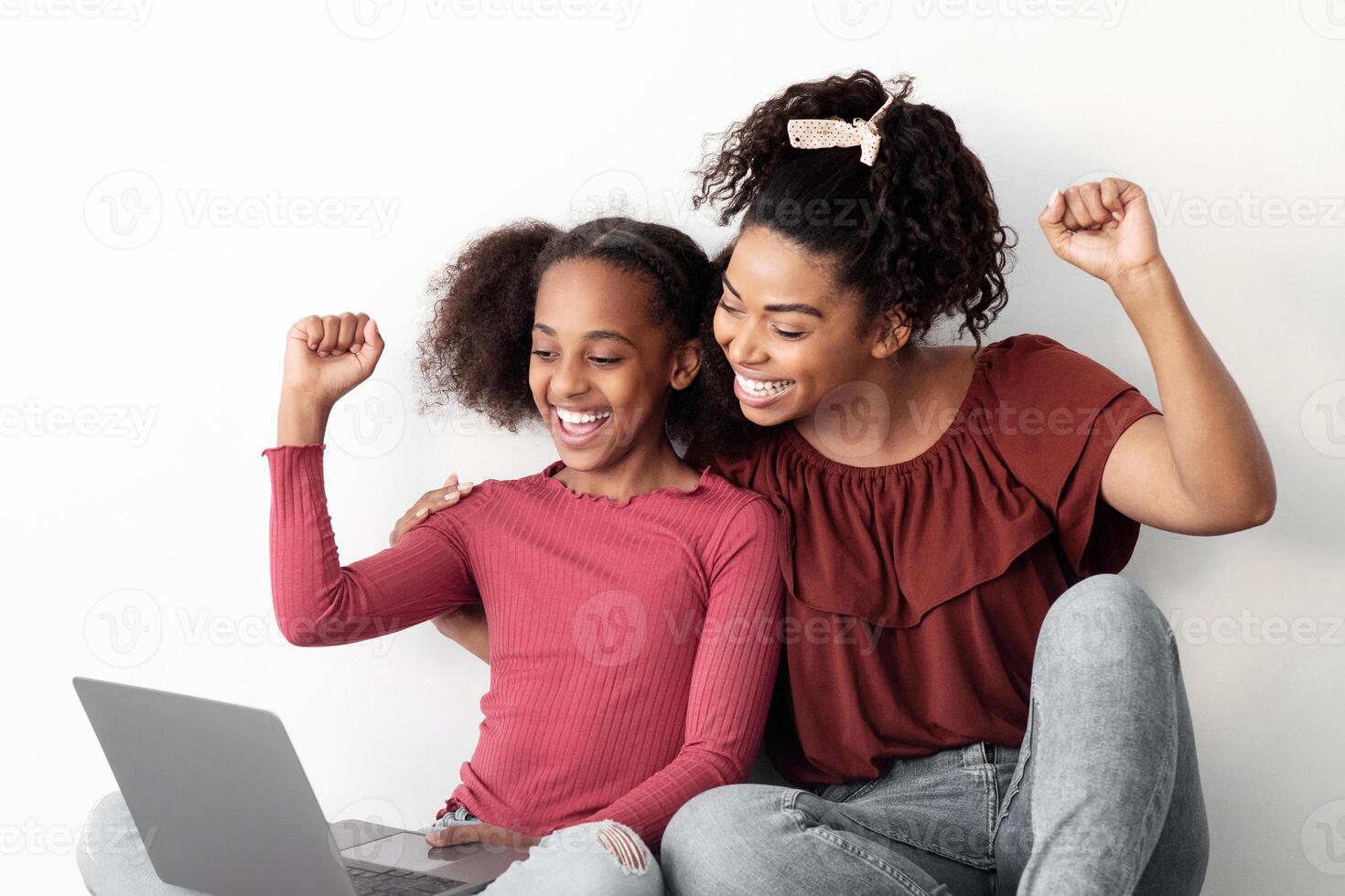 Emotional african american mom and kid gaming online together, using laptop, sitting on floor by white empty wall, raising hands up and smiling, playing games, panorama with copy space photo