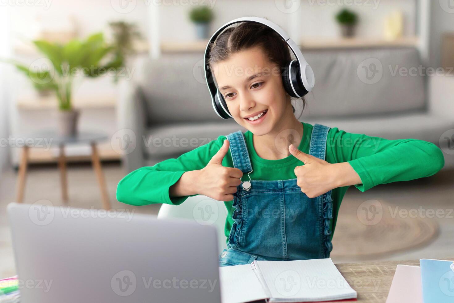 Smiling girl in headphones enjoying elearning sitting at table showing thumbs up to pc webcam, recommending e-study and online school for children, using modern tech. Clever kid, homeschooling concept photo