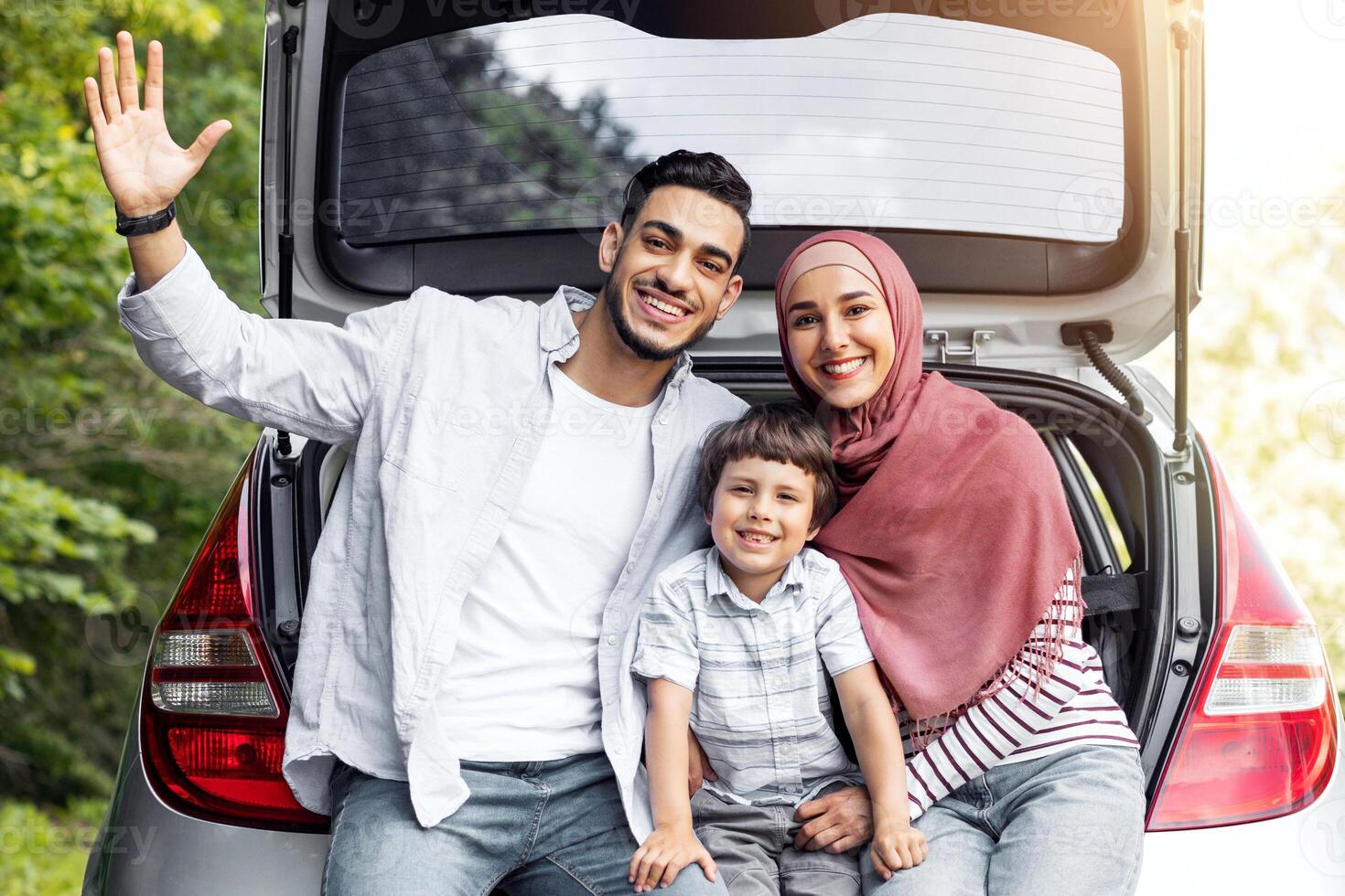 Family travel at own new auto. Satisfied millennial islamic male and female in hijab with son, sitting in car trunk and waving hand, resting from trip together. Vacation, rest from trip and emotions photo