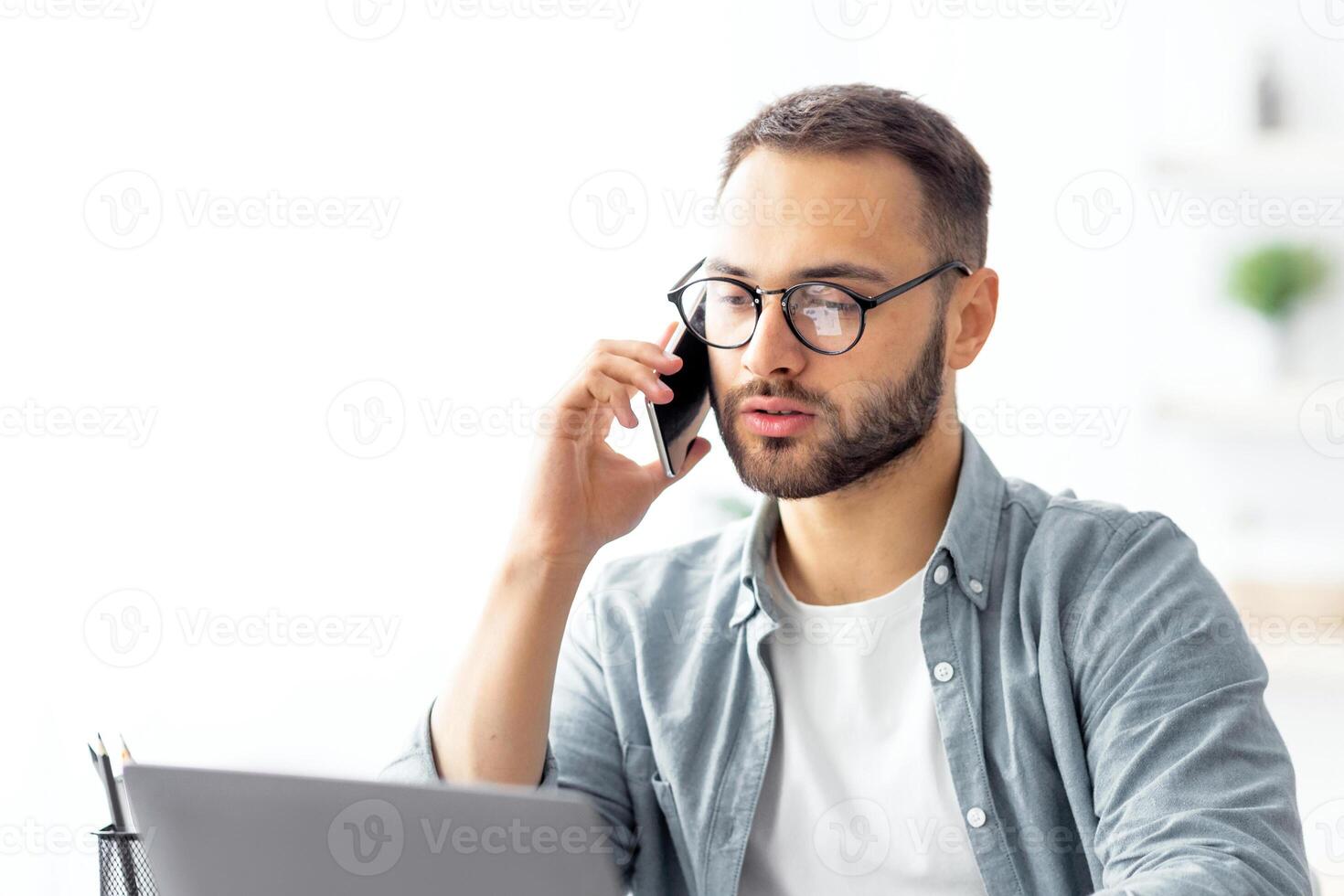 Portrait of happy young guy using laptop, speaking on cellphone, having freelance job at home. Millennial man communicating remotely, having phone call during online business meeting photo