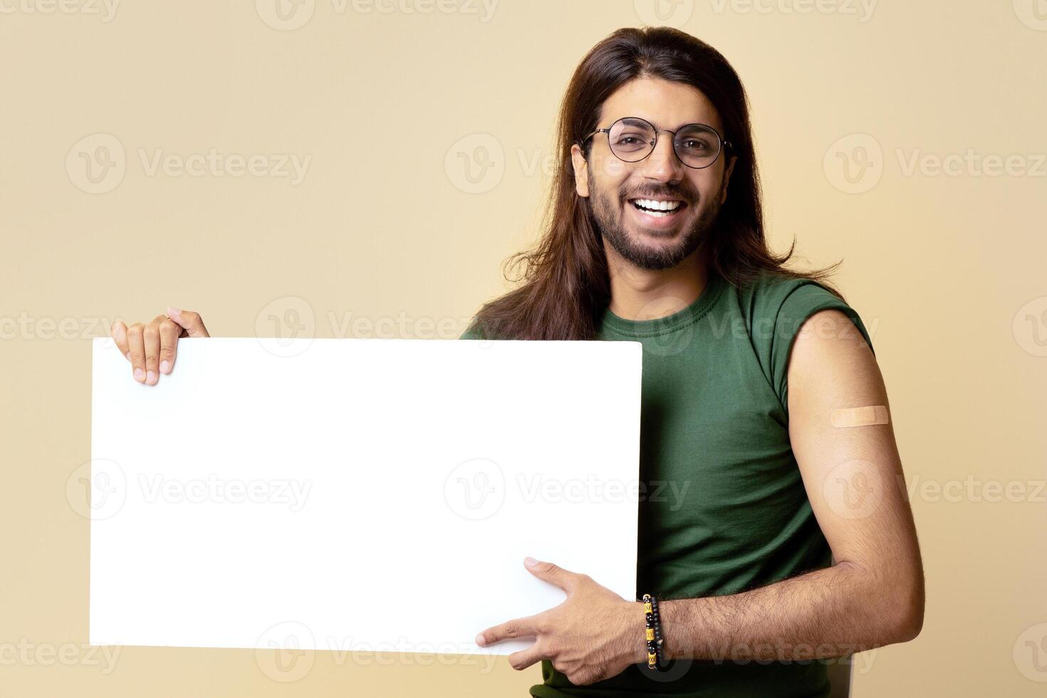 Happy vaccinated young indian man with medical band on shoulder sitting on chair over beige studio background and showing white empty placard for text or advertisement, smiling at camera photo