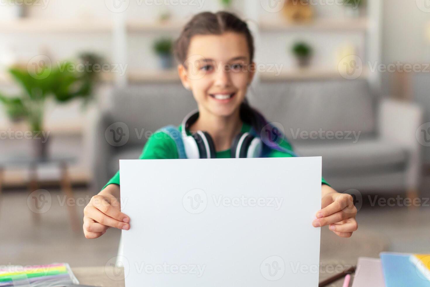 Portrait of smiling teenage girl holding and showing blank white sheet of paper, sitting at desk in living room, selective focus, blurred background. Free copy space for text photo