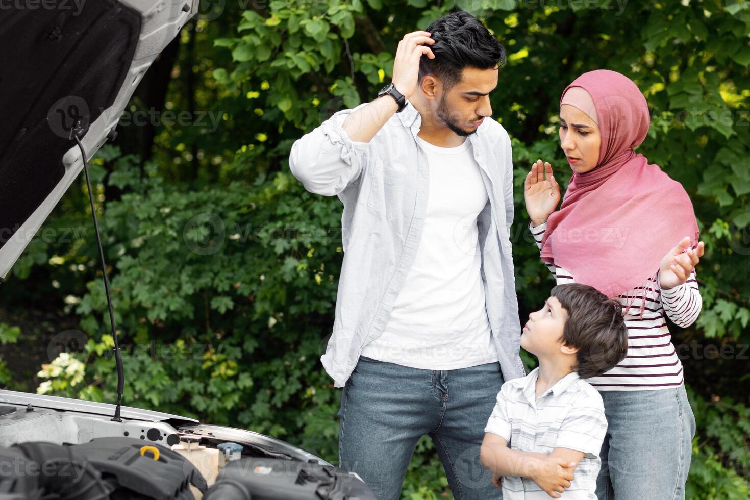 Sad disappointed millennial islamic husband and wife in hijab with child stand near broken car with open hood, outdoor. Family travel, vacation together and trip, people emotion and incident at road photo