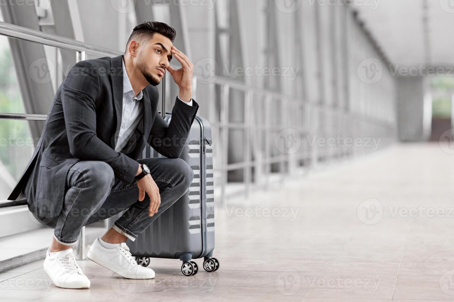 Missed Flight. Portrait Of Upset Middle Eastern Man Sitting Next To Suitcase At Airport, Pensive Young Arab Guy Waiting With Luggage At Terminal Corridor And Touching Forehead, Copy Space photo