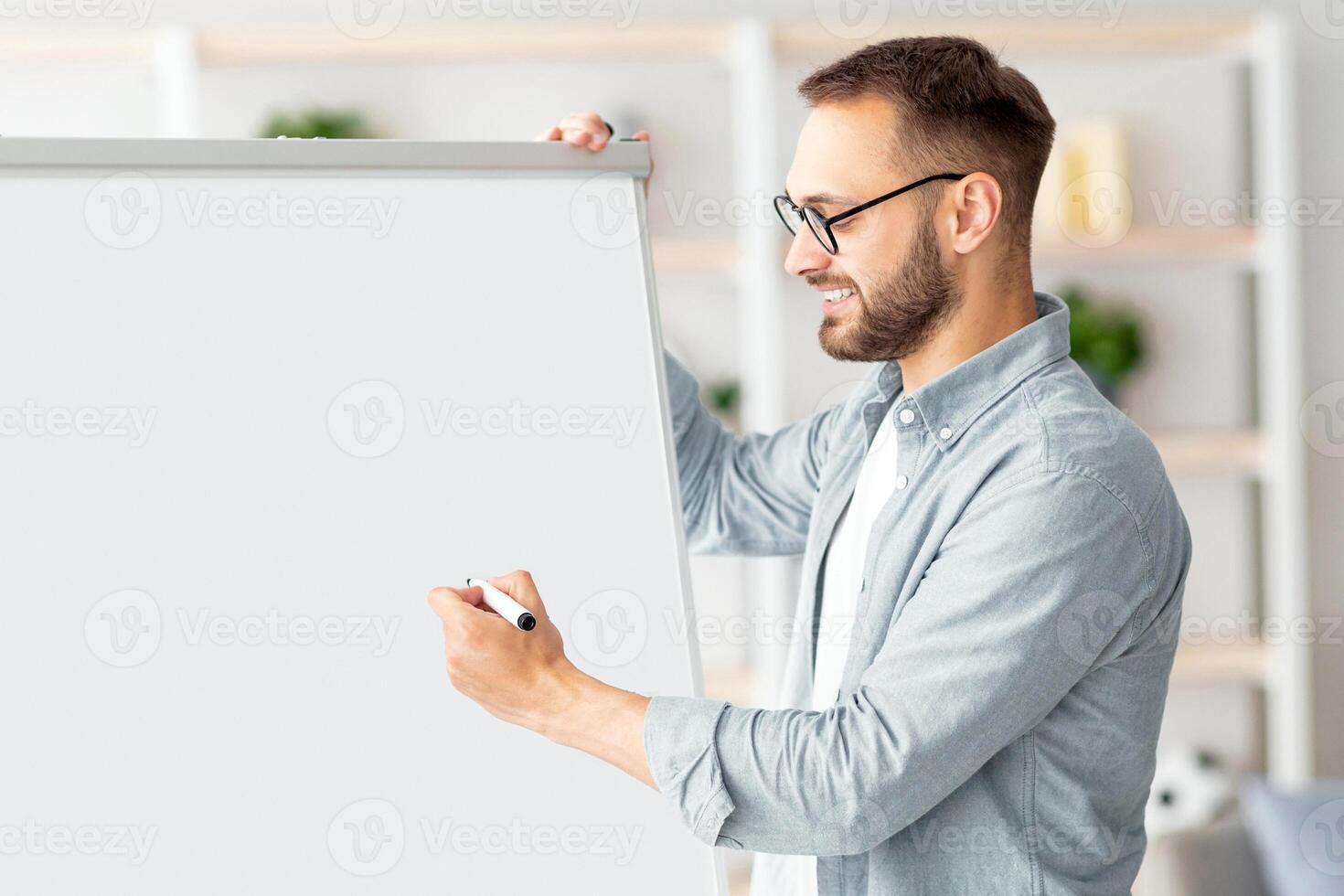 Young Caucasian male teacher giving online class, explaining English rules, writing on blackboard at home office. Remote tutoring and distance education during coronavirus lockdown concept photo