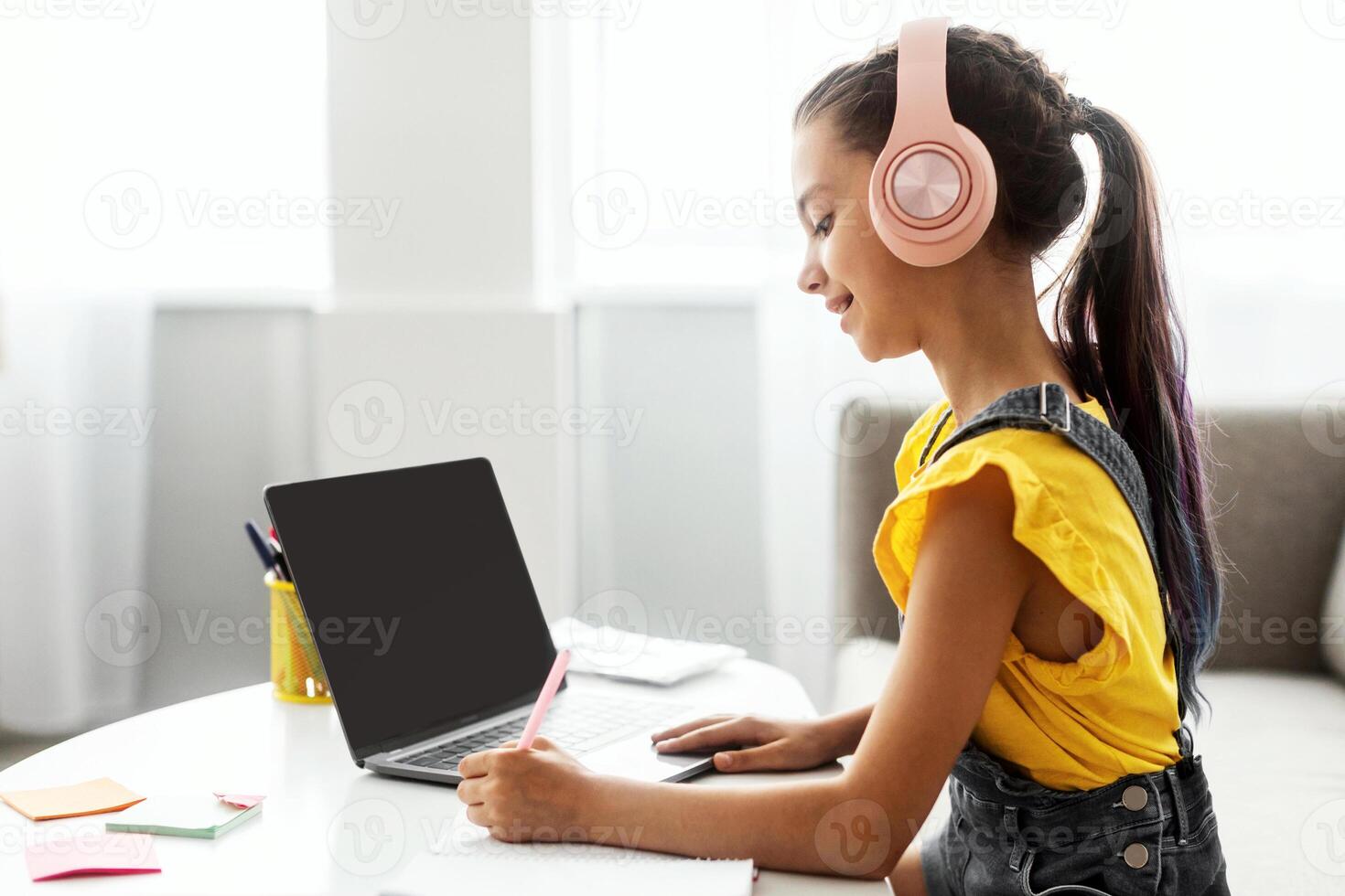 Side view profile portrait of girl in pink headset doing homework, sitting on couch at table using laptop with empty screen, writing essay or making test, taking notes in textbook, free copy space photo