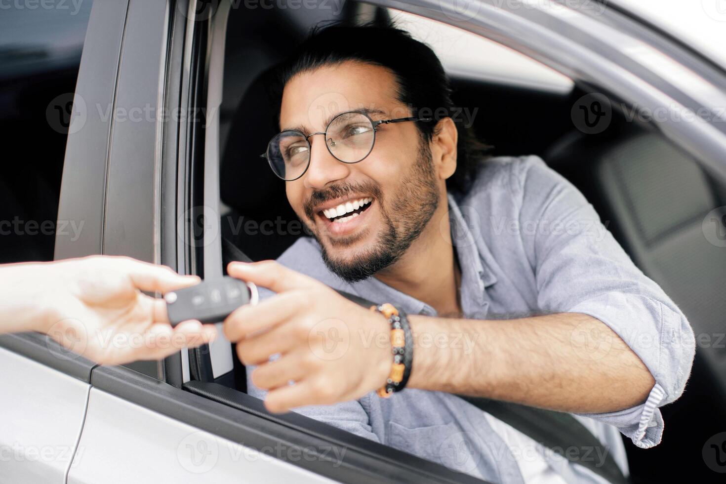 Handsome middle-eastern young man sitting inside nice automobile and taking car key from leasing or renting service person. Cheerful arab guy doing test-drive before buying new automobile photo