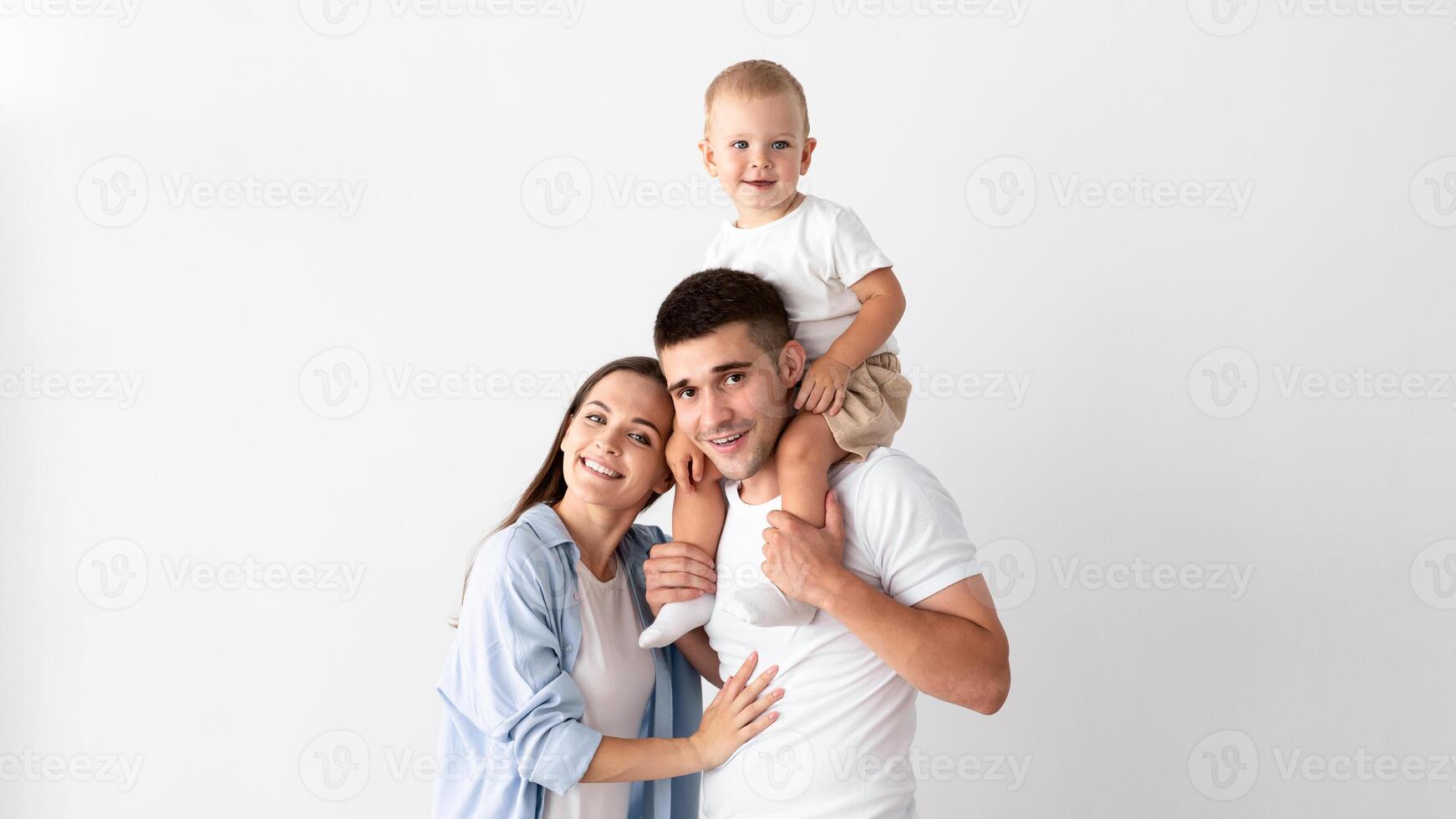 Portrait Of Happy Young Mother, Father And Their Cute Toddler Son Posing Together Over White Wall At Home, Cheerful Parents And Adorable Infant Child Embracing And Smiling At Camera, Copy Space photo