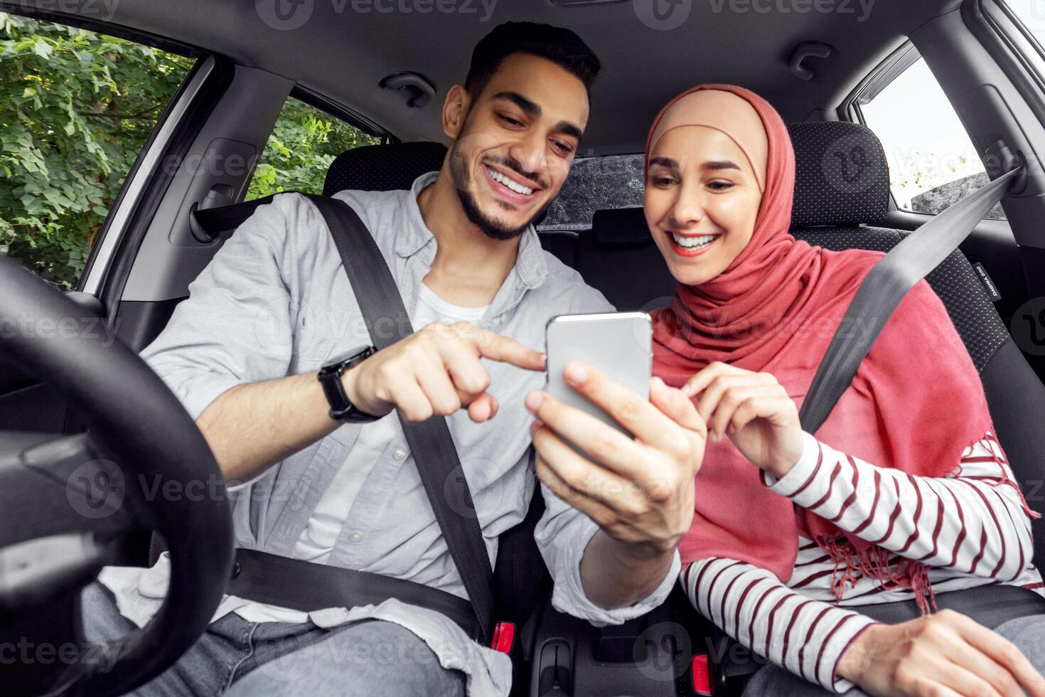 Smiling young arab woman in hijab and guy looking for way on map on smartphone while driving in car, outdoor, panorama. Travel app, great mood, new car purchase, weekend trip and journey together photo