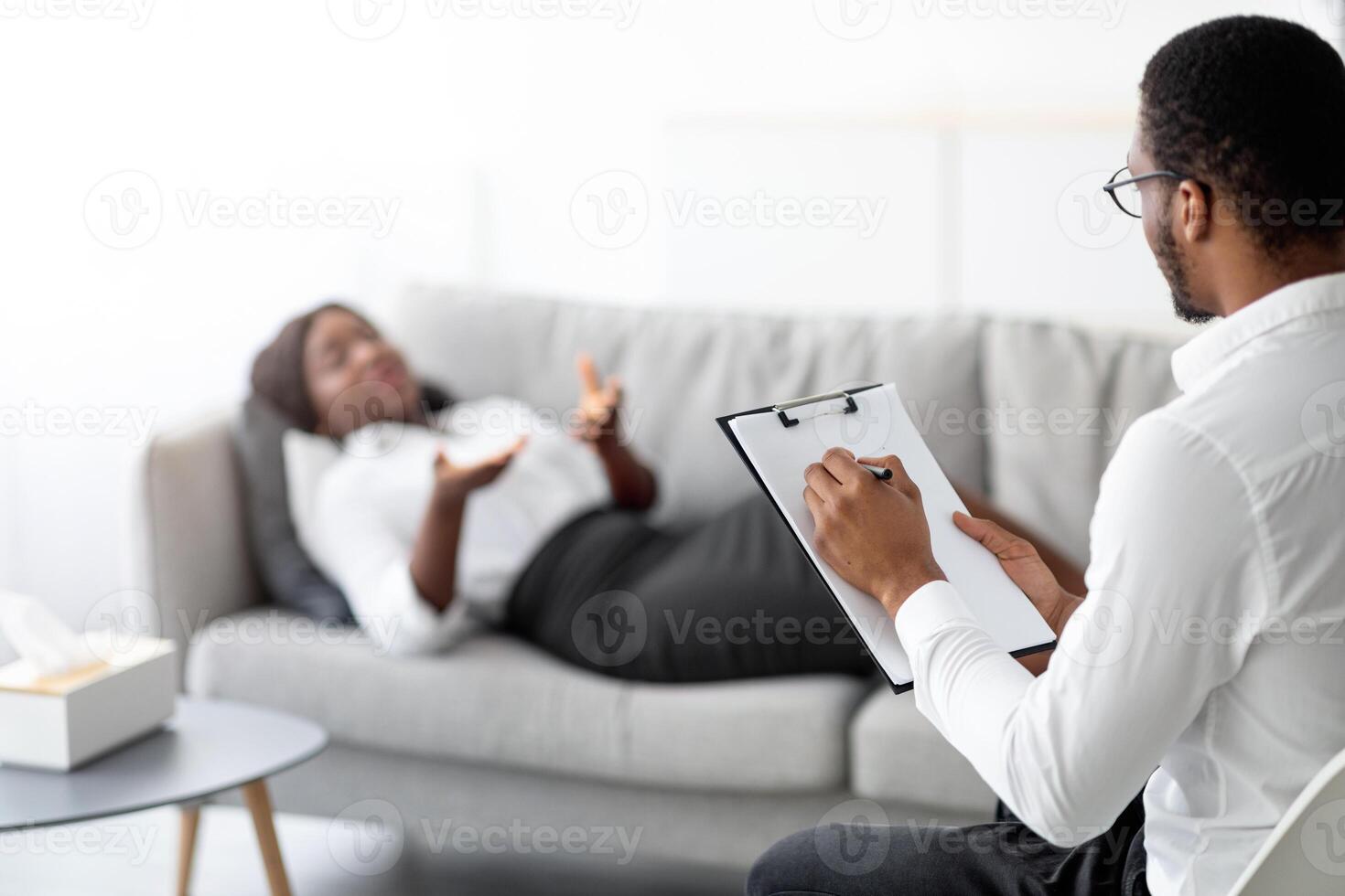 Young black woman on consultation with psychologist, lying on sofa at office, explaining about her problem to doctor, selective focus. Millennial lady speaking to psychotherapist at clinic, copy space photo
