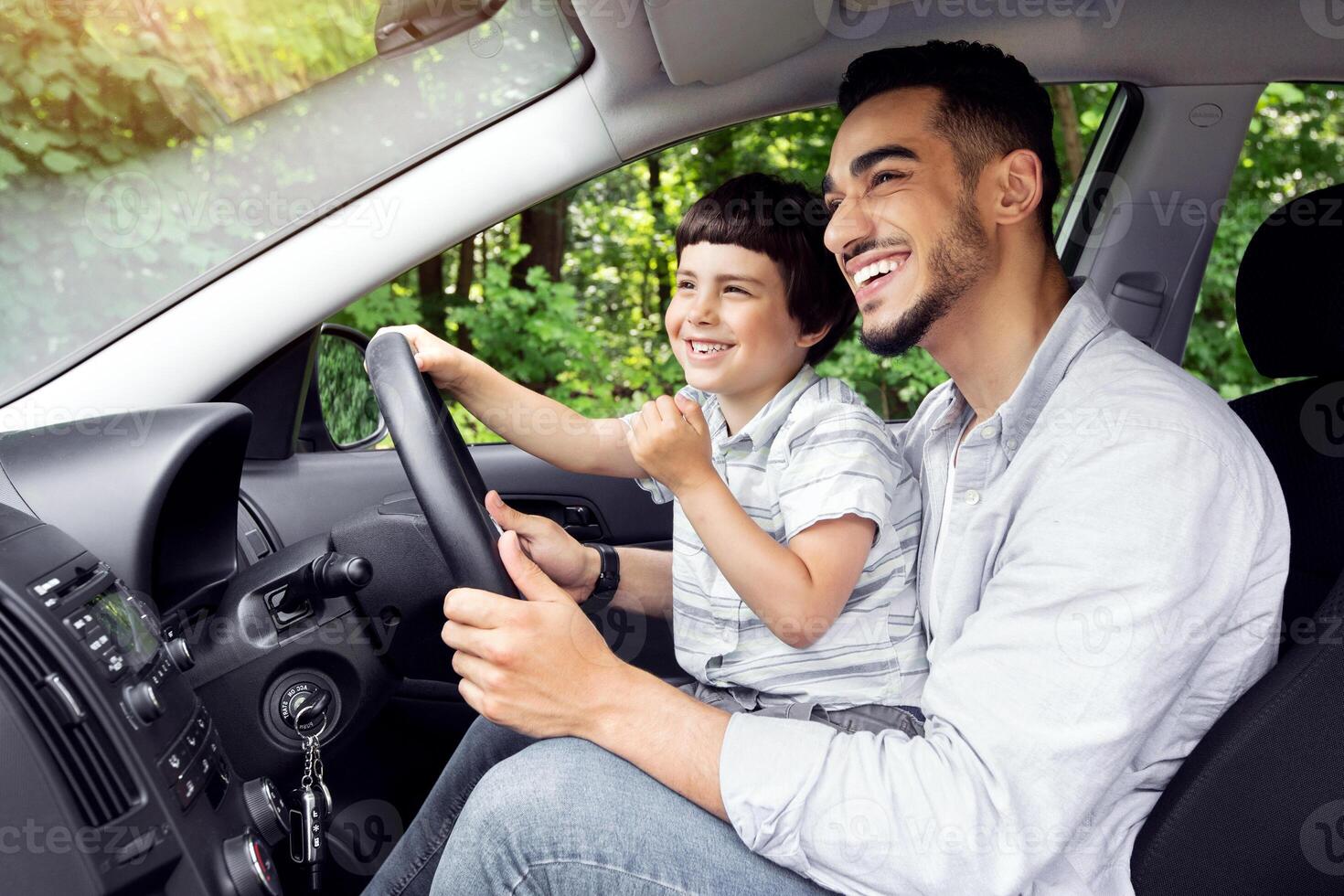 Young Arab Father Giving Driving Lessons To His Little Son, Happy Preteen Boy Sitting On Dad's Lap In Modern Car And Holding Steering Wheel, Enjoying Road Trip And Spending Time Together, Side View photo