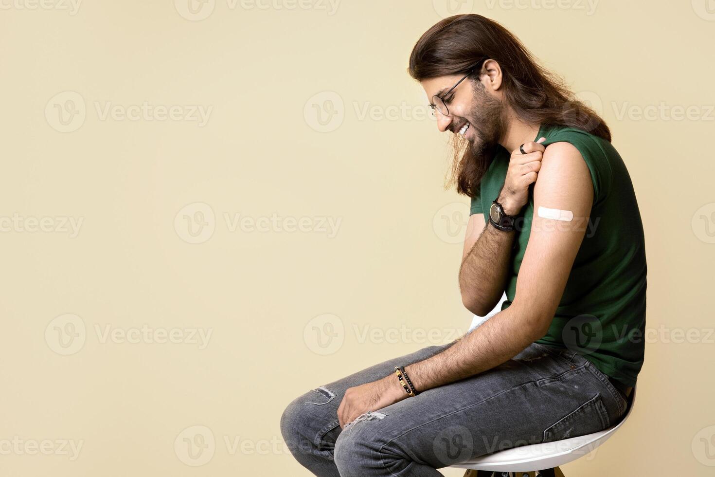 Relaxed young arab guy with adhesive medical band on shoulder resting on chair after immunization, beige studio background, panorama with copy space. Vaccination while COVID-19 pandemic concept photo