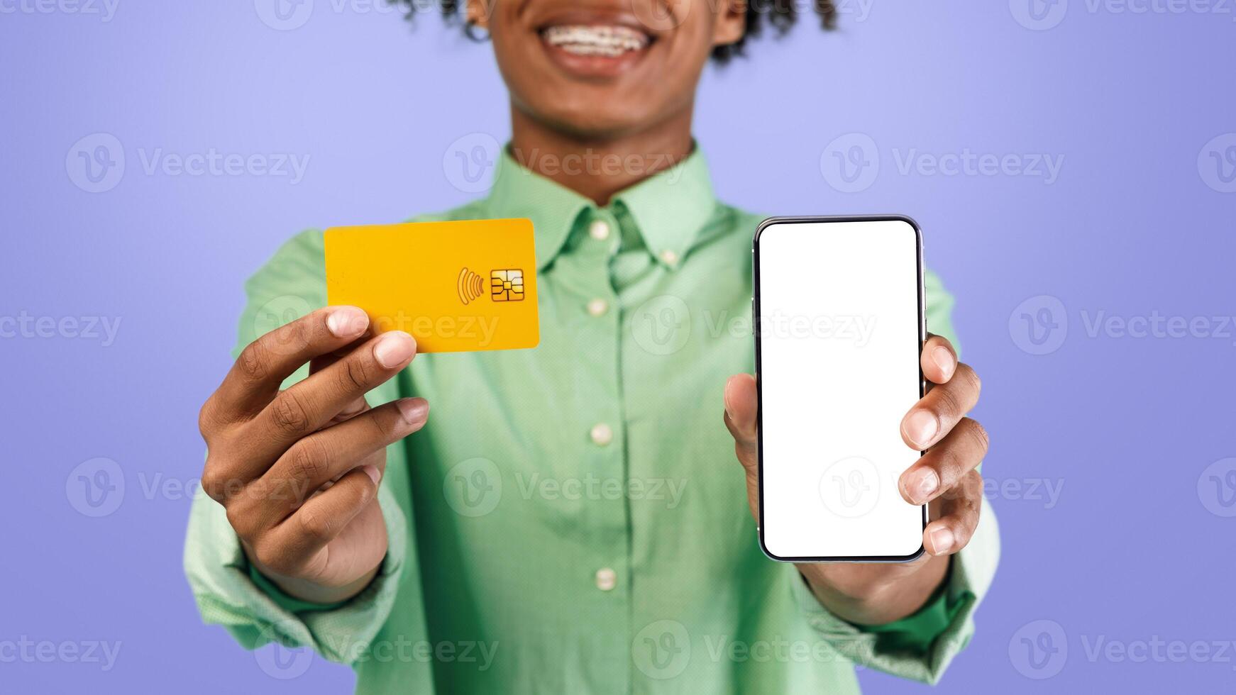 Cool mobile shopping app. Excited young black guy holding credit card and showing cellphone with blank screen to camera. Gadget with copy space for mockup, digital pay photo