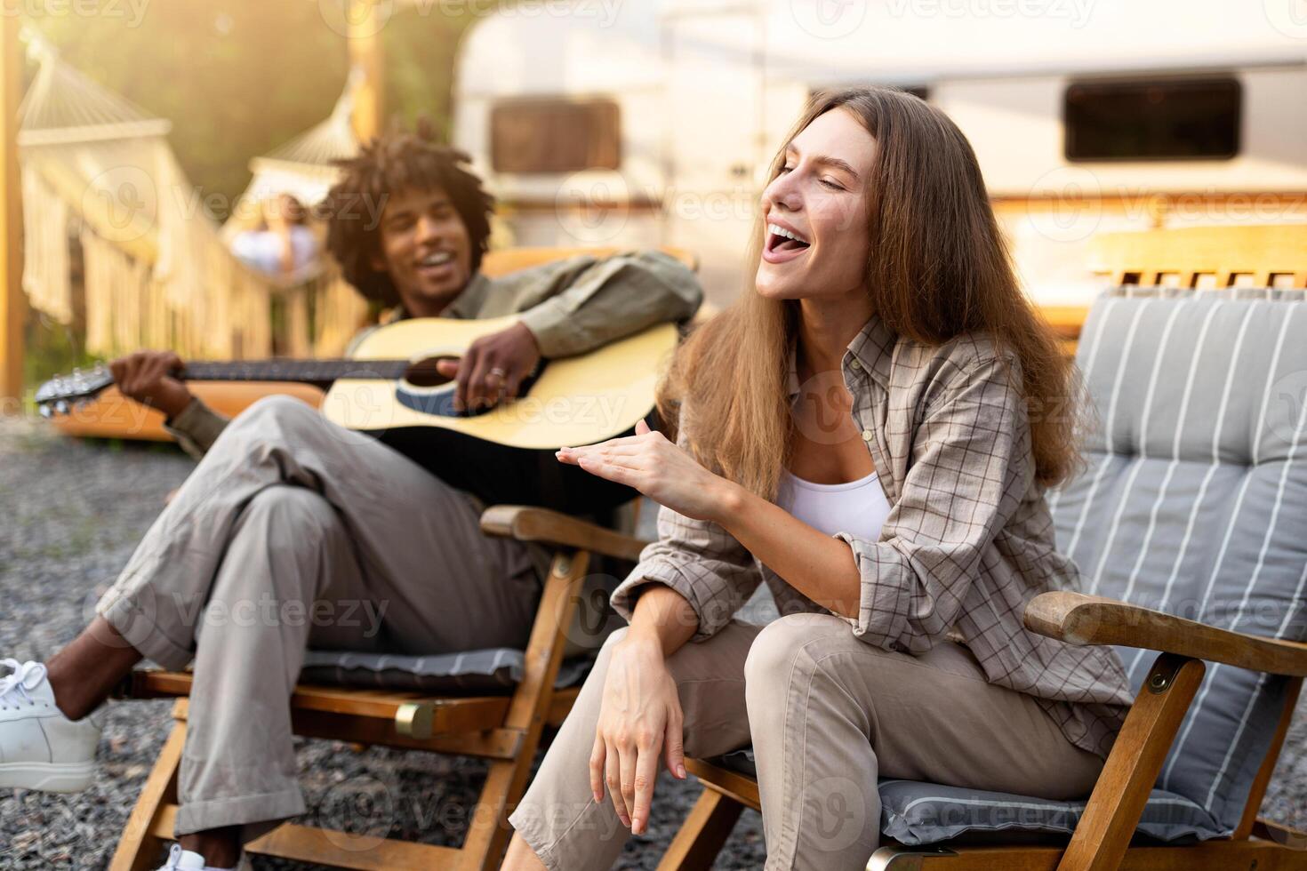 Carefree diverse friends singing and playing guitar, enjoying music near RV on camping trip. Young lady and black guy having fun on summer holidays, spending time in nature photo