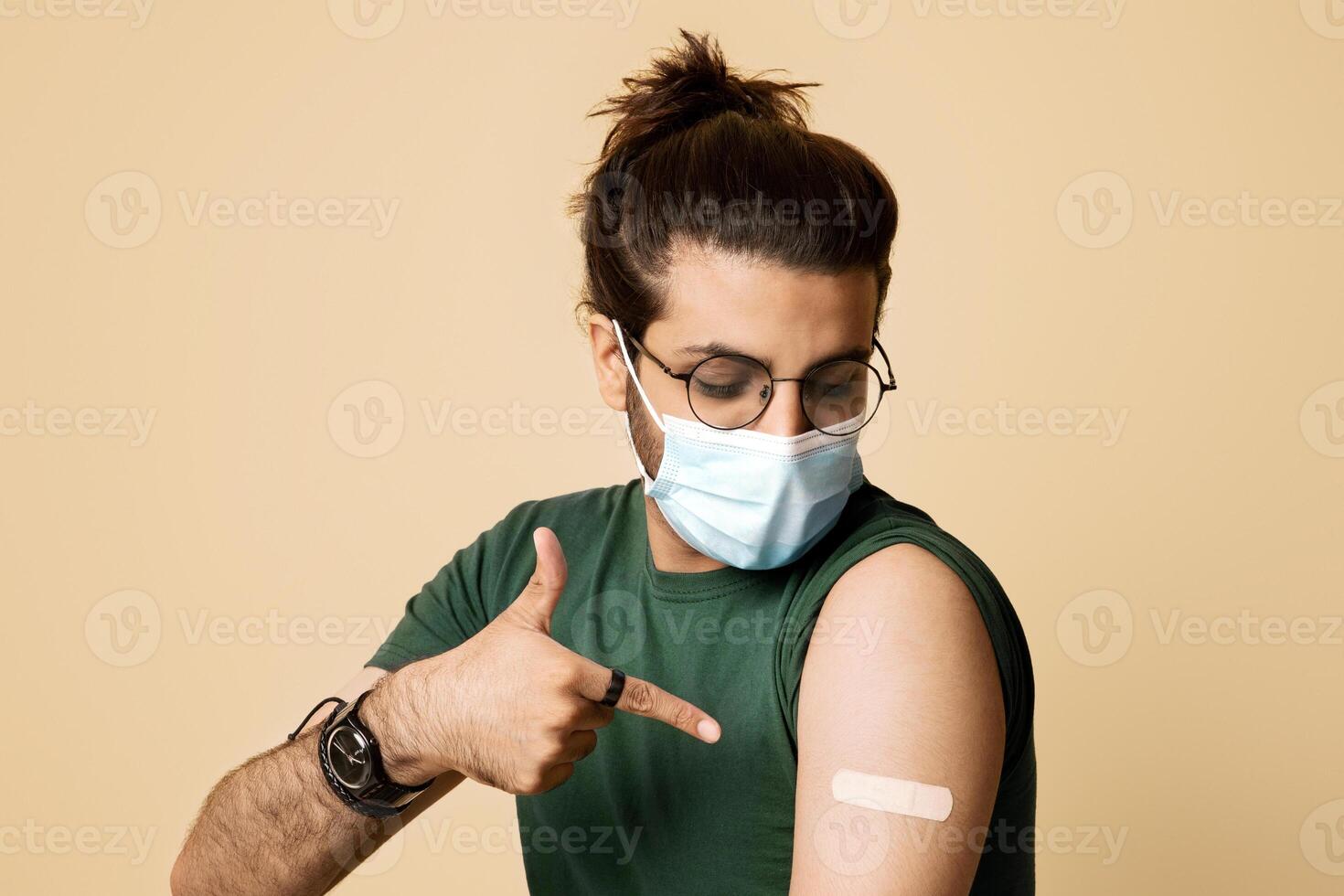 Long-haired indian guy in protective face mask sitting on chair over studio background, pointing at medical band on his shoulder, young middle-eastern man got vaccinated against COVID-19, copy space photo