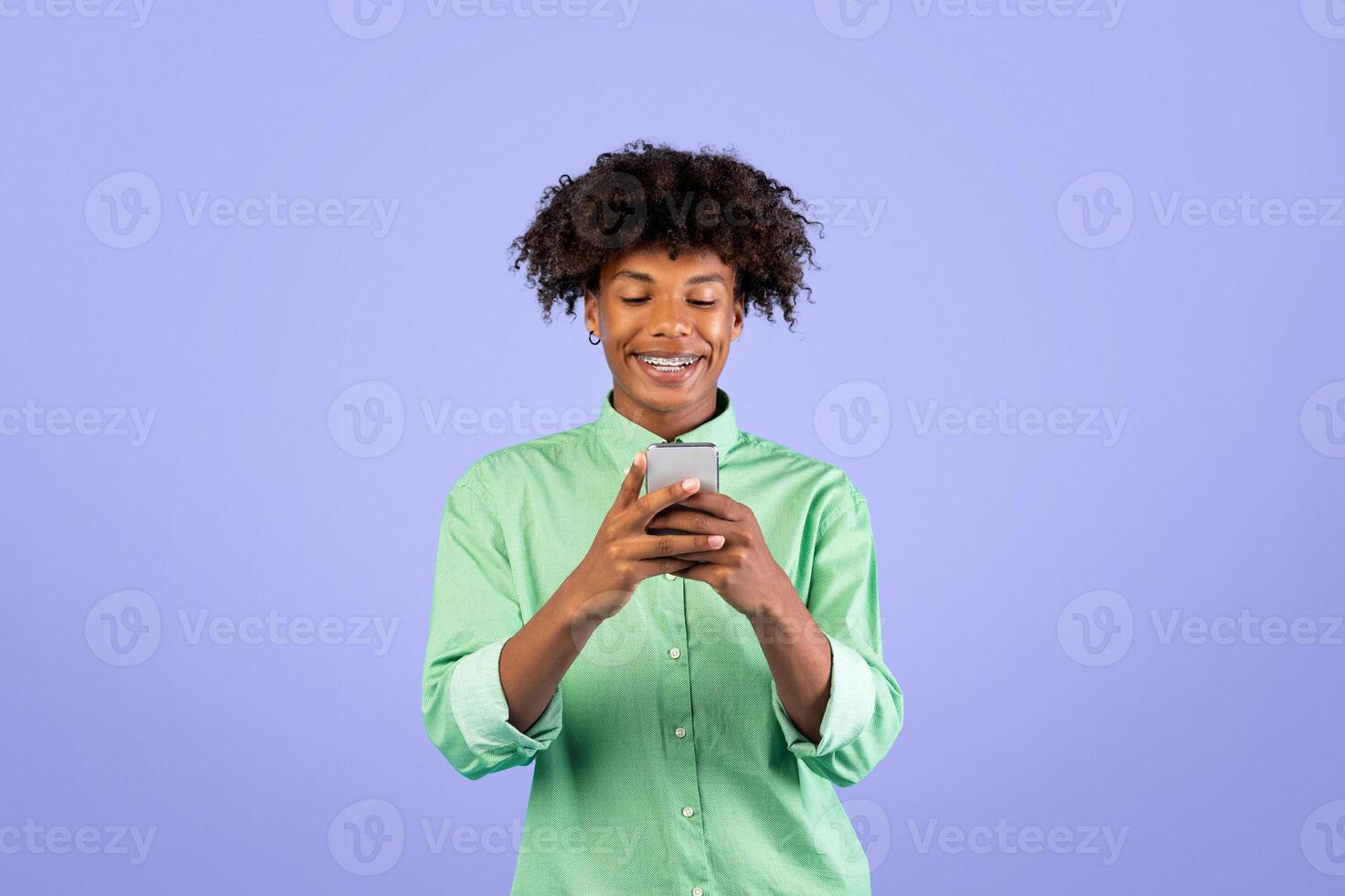 People and gadgets. Happy black guy using smartphone, smiling, browsing internet and scrolling social media news feed standing over blue studio background photo