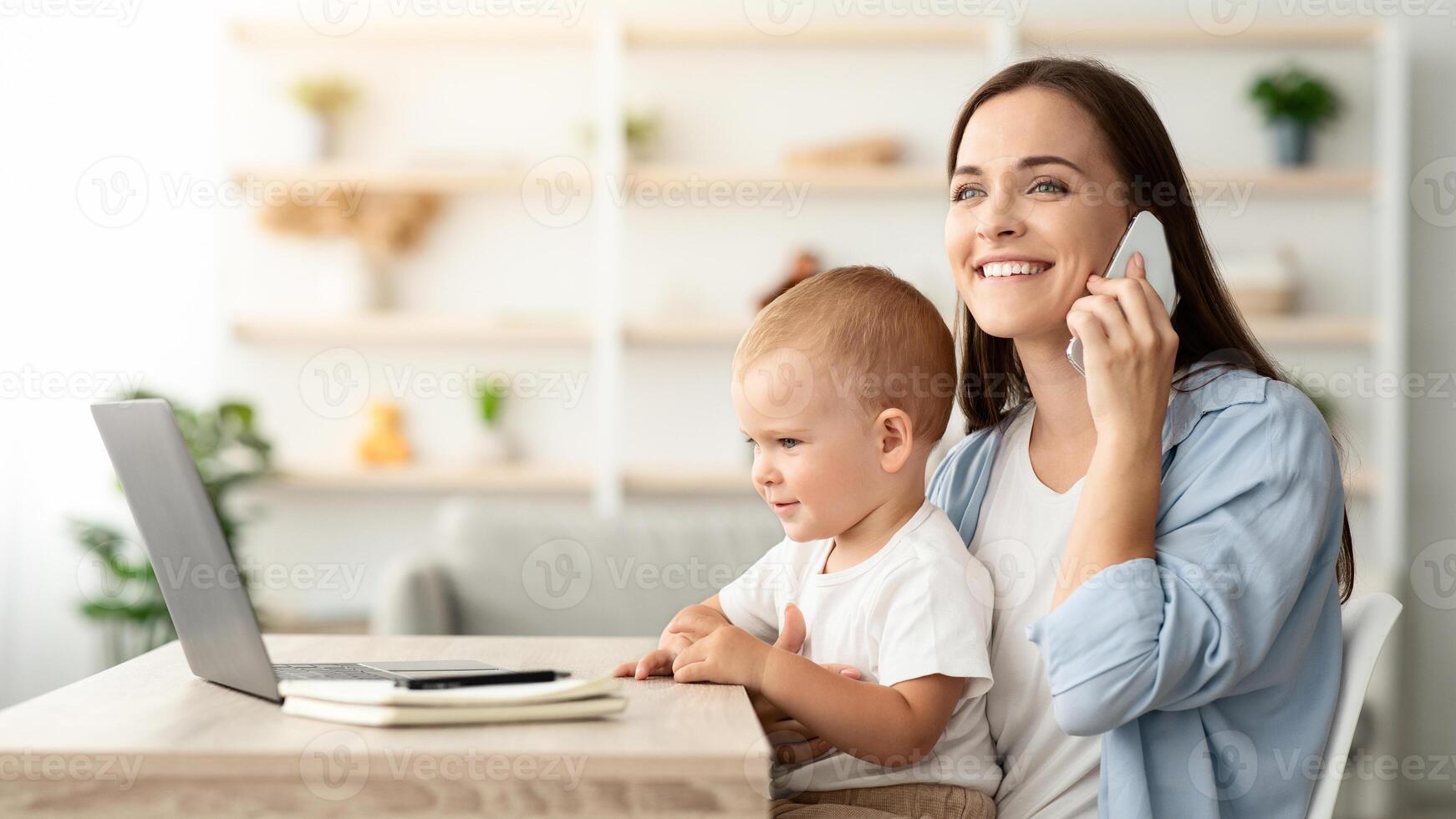 Smiling Young Mother Holding Little Baby And Talking On Cellphone At Home, Happy Freelancer Lady Sitting At Desk With Laptop Computer, Working Remotely And Taking Care About Toddler Son, Copy Space photo