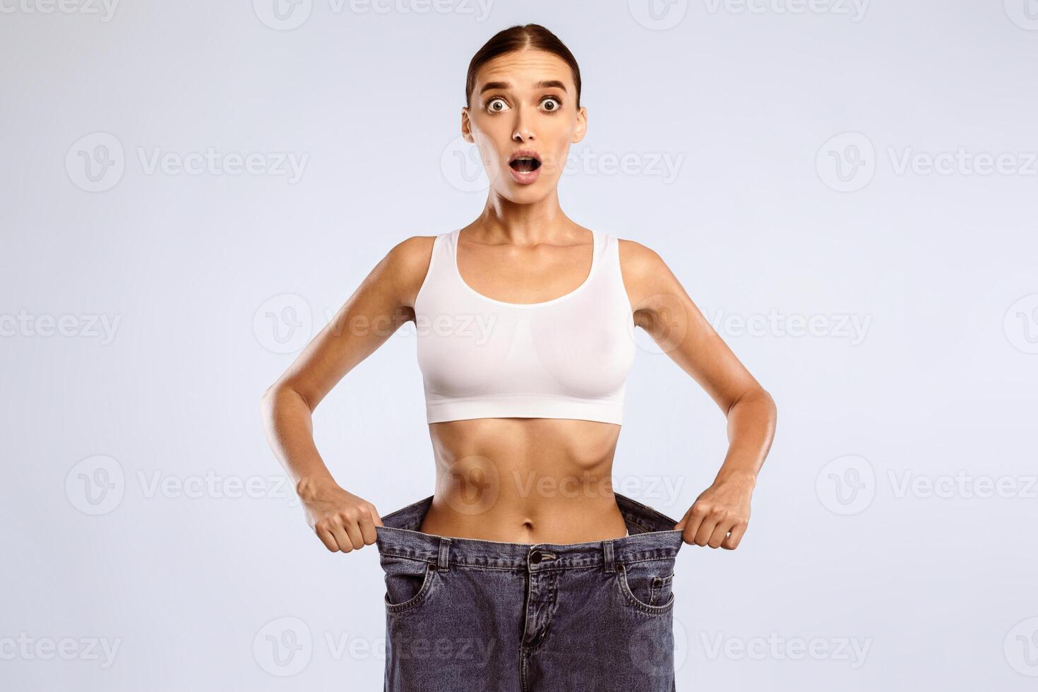 Wow. Portrait Of Shocked Suprised Young Woman Wearing Too Big Loose Jeans After Successful Weight Loss, Posing and Looking At Camera With Open Mouth In Amazement Standing Over Blue Studio Background photo