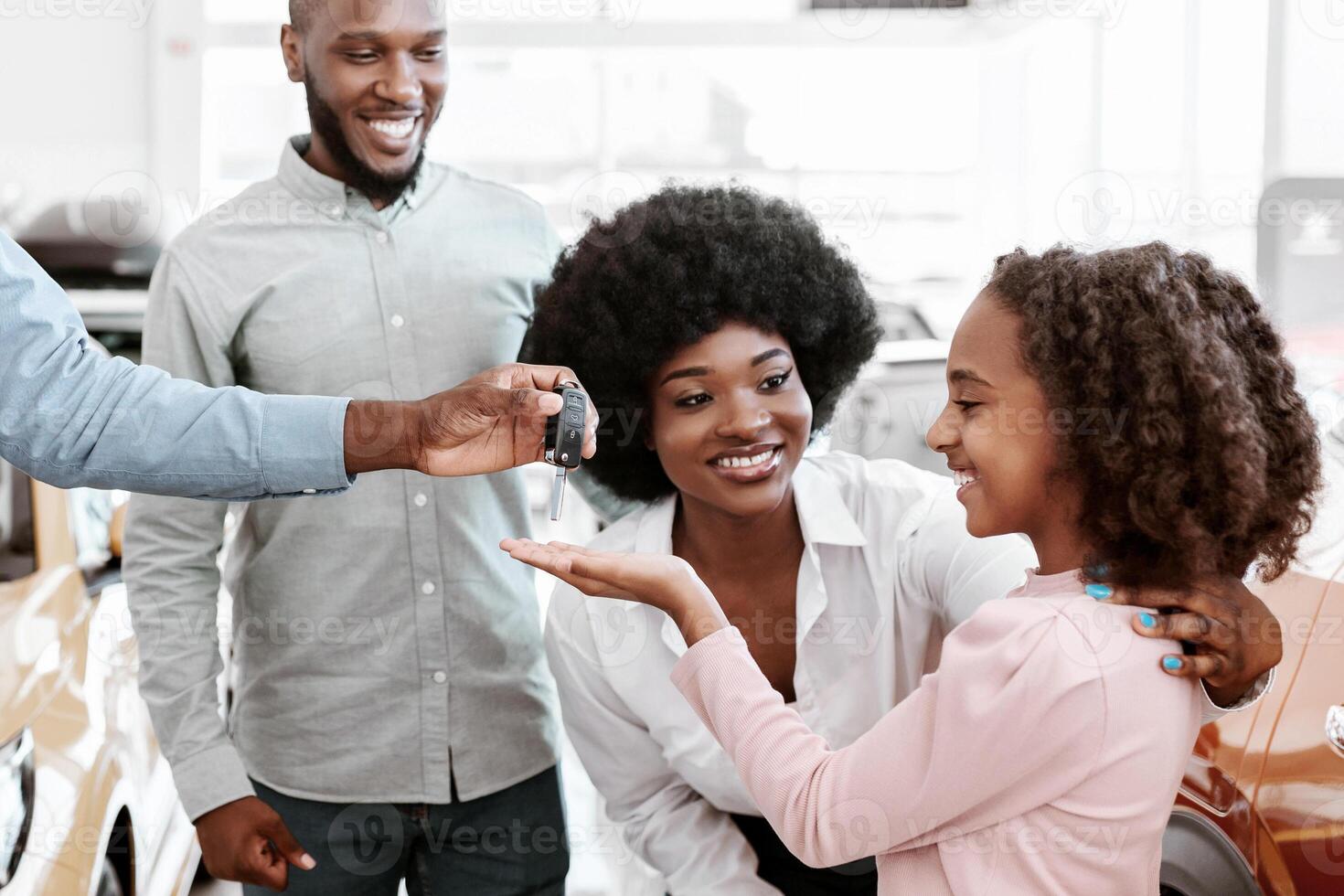 Positive auto salesman giving car key to cute black girl and her parents at dealership store. Cheerful African American family shopping for new vehicle at automotive showroom photo