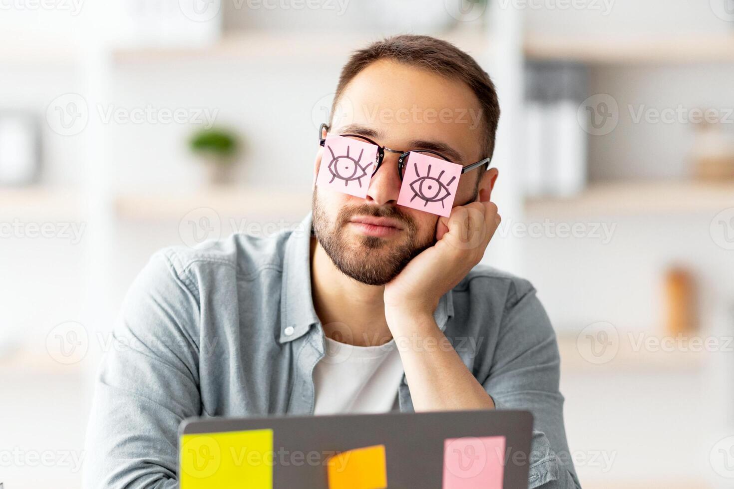 Workaholism, exhaustion at workplace concept. Tired young man having stickers with open eyes drawing on glasses, sleeping near laptop, pretending to work at home office photo
