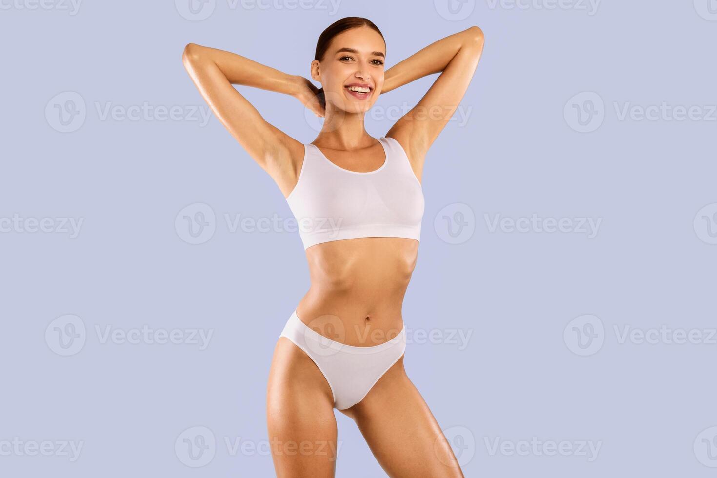Portrait of happy smiling fit woman wearing lingerie posing isolated om blue studio background, raising arms up. Female with flat belly in white underwear top bra and panties showing smooth armpits photo