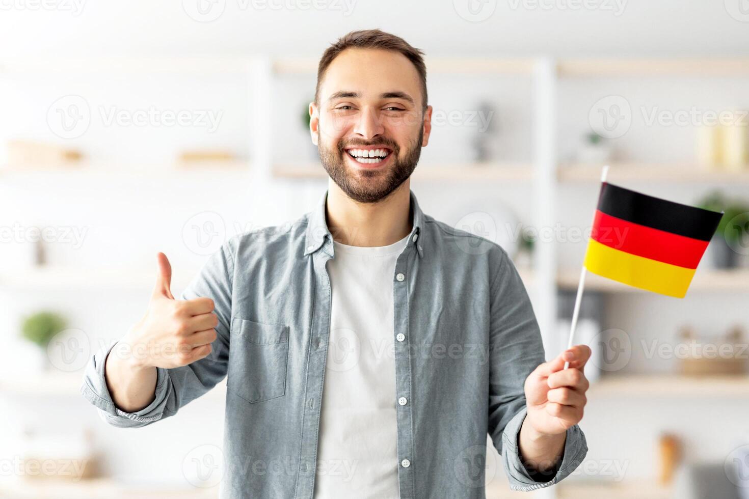 Happy Caucasian man showing thumb up and flag of Germany, posing and smiling at camera indoors. Cheerful millennial student recommending foreign education, learning German language photo