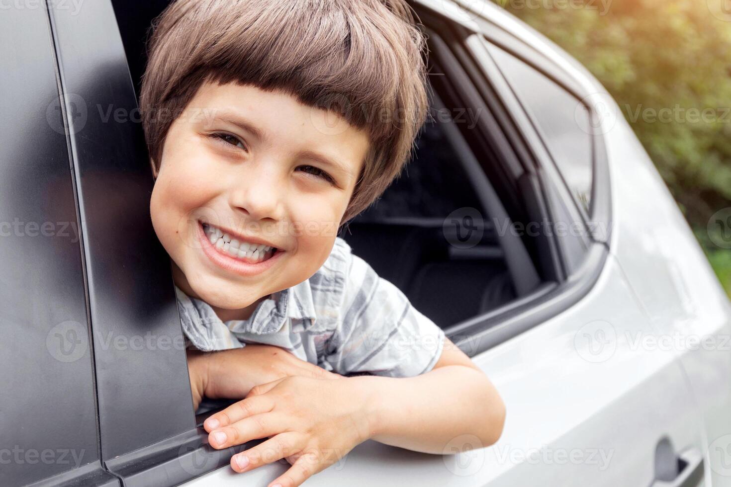 Glad little kid looks out the open window and enjoys trip with his parents in car, outdoors in summer, copy space. Weekend with family, vacation together, road and journey by auto, facial expression photo