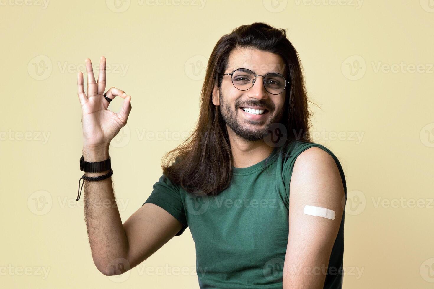Cheerful long-haired indian guy showing okay gesture and smiling at camera, got vaccinated against COVID-19, having adhesive medical band on shoulder, beige studio background, copy space photo