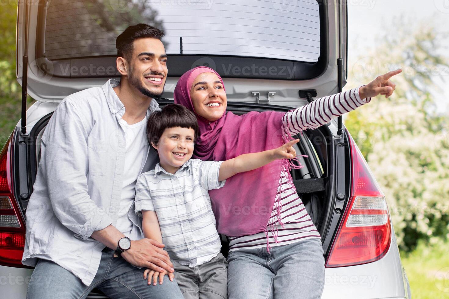 Happy Muslim Family With Little Son Sitting In Car Trunk Outdoors, Excited Middle Eastern Parents And Little Male Child Pointing Aside With Hand, Enjoying Road Trip With Their New Automobile Together photo