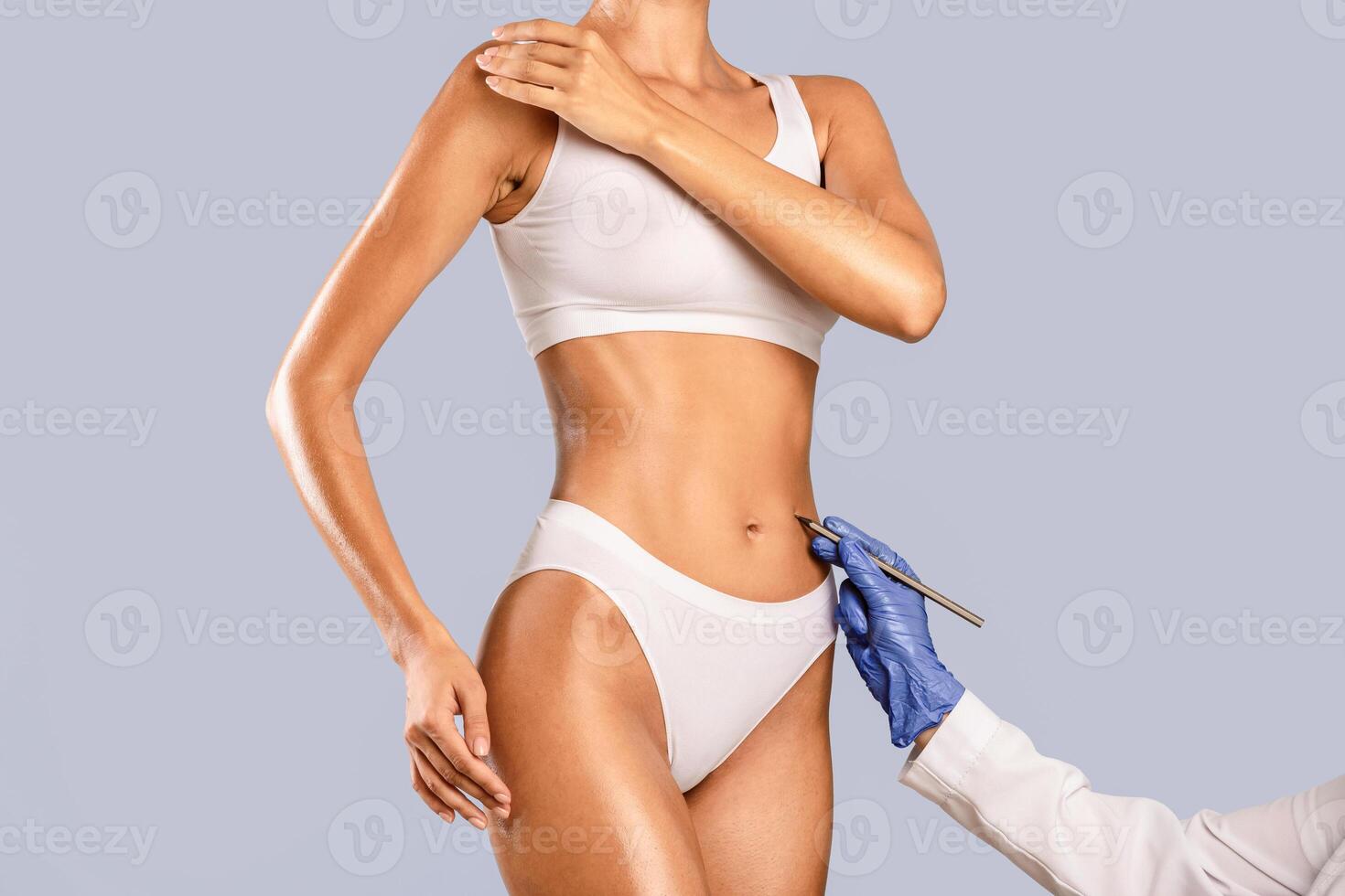 Doctor drawing marks on female stomach, preparing for cosmetic surgery operation isolated on blue studio background. Young lady getting ready for aestetic correction procedure, mesotherapy, lipolysis photo