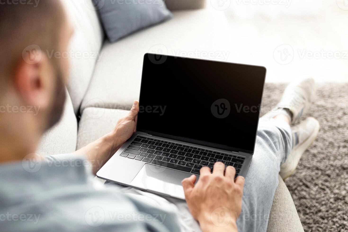 Unrecognizable young man using laptop on comfy sofa at home, mockup for website design on screen. Millennial guy holding portable pc with copy space, working or studying online photo