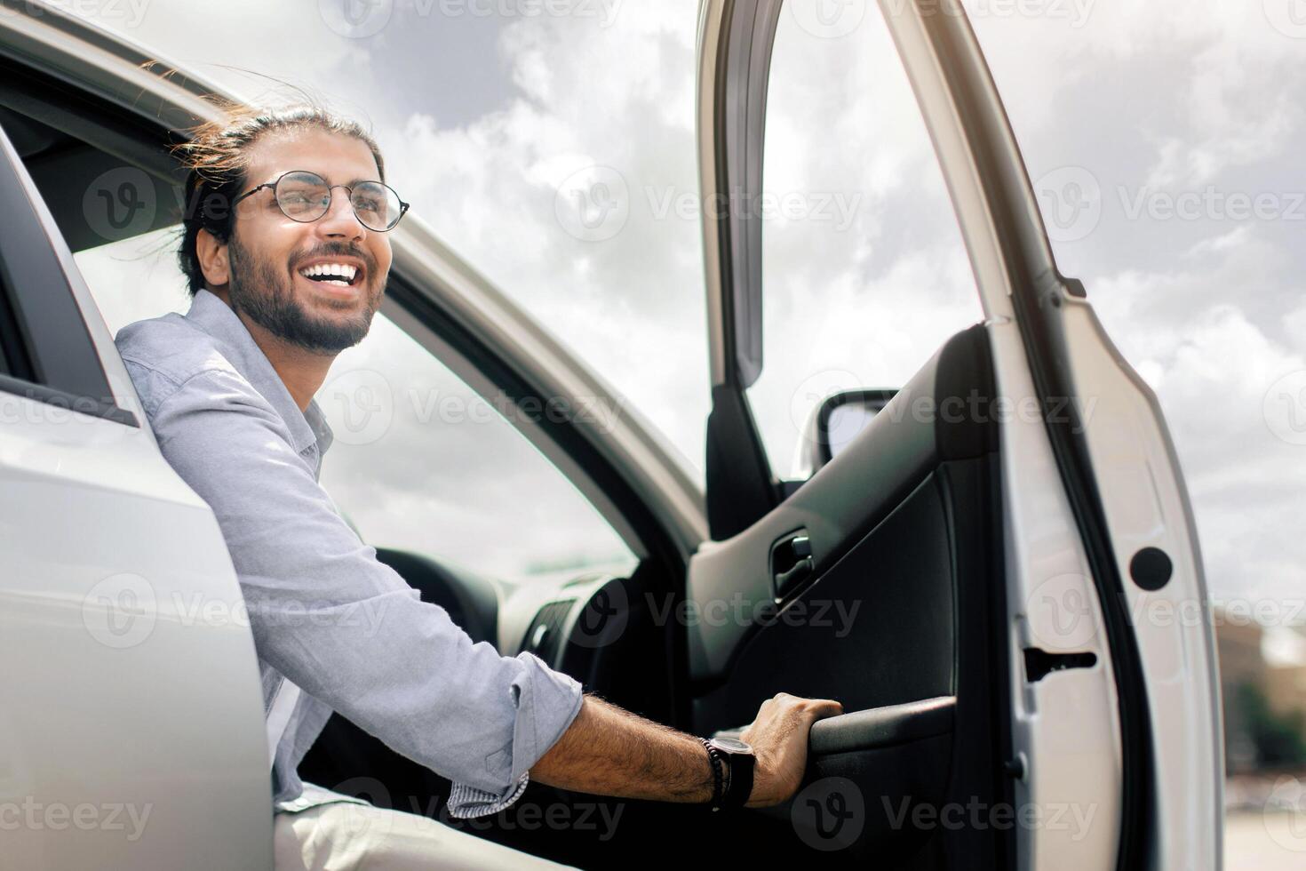 Positive young middle-eastern man going out the car, low angle photo, copy space. Happy arab guy open automobile door and smiling, getting in. Auto test-drive, leasing, buying, renting concept photo