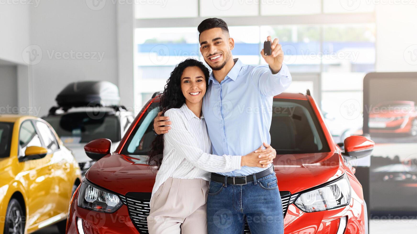 Happy beautiful loving muslim family young arab man and long-haired woman buying new car in salon, standing over red luxury automobile, hugging and showing key from their new auto, copy space photo