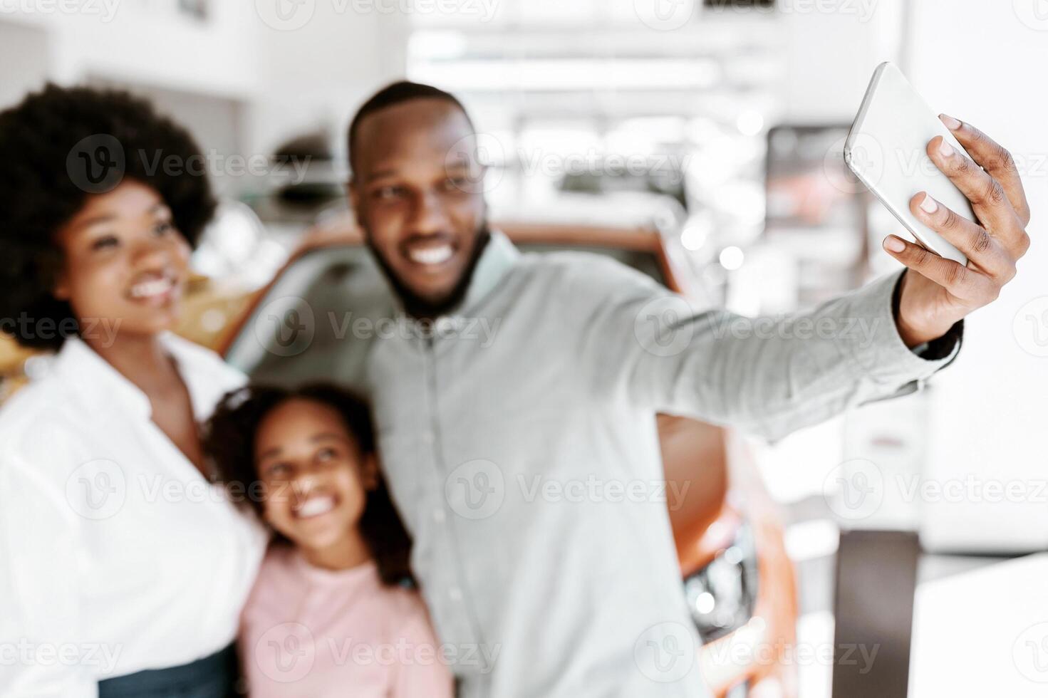 Joyful black family taking smartphone selfie near their new luxury car at auto dealership, selective focus. Positive Afro parents and child making photo at automobile showroom, copy space