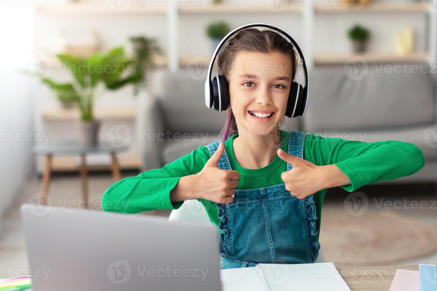 Distant Education. Portrait Of Cheerful Teen Girl In Headphones Using Laptop In Living Room, Sitting At Table And Showing Thumb Up Gesture At Camera, Female Pupil Studying Online With Computer photo