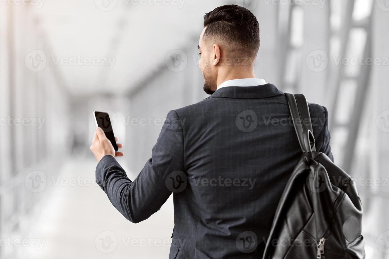 Booking Hotel Online. Arab Man With Backpack Using Smartphone With Blank Black Screen While Walking In Airport Terminal, Male Traveller Enjoying Modern Technologies, Rear View With Free Space, Mockup photo