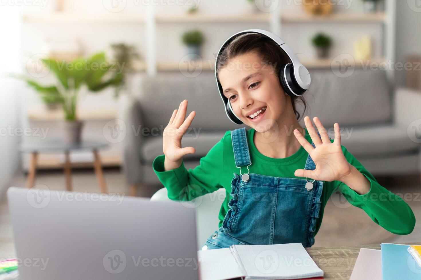 Distance Education. Portrait of smiling schoolgirl making call with teacher and classmates, using pc. Cheerful student in wireless headphones talking and waving both hands showing ten 10 fingers photo