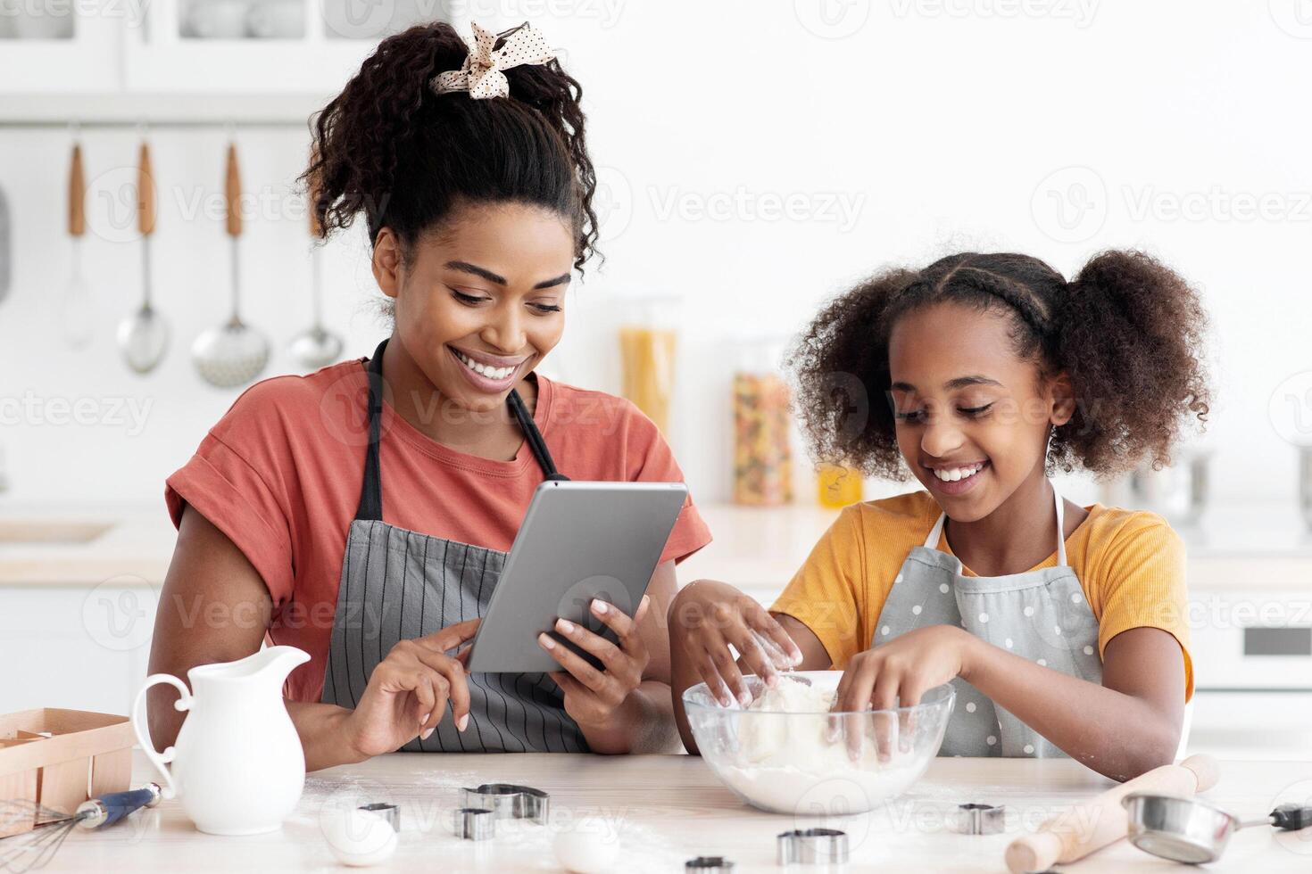 Happy black mother and daughter baking together at home, reading culinary blog on Internet, using digital tablet, african american mom and teen kid making dough for cookies or cake photo