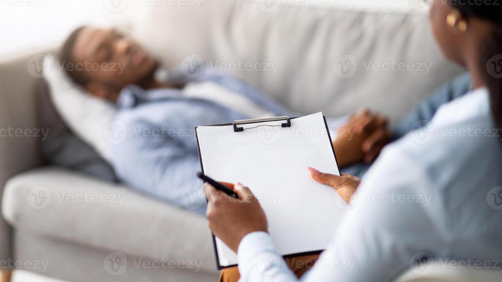 Stress and psychological trauma. Psychologist working with depressed black man at office, mockup on clipboard. African American male patient lying on couch while having session with therapist photo