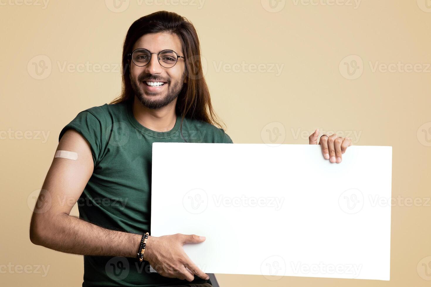Healthy arab guy got vaccinated against coronavirus, sitting on chair over beige studio background, showing adhesive plaster on his shoulder and empty horizontal board for text or advertisement photo