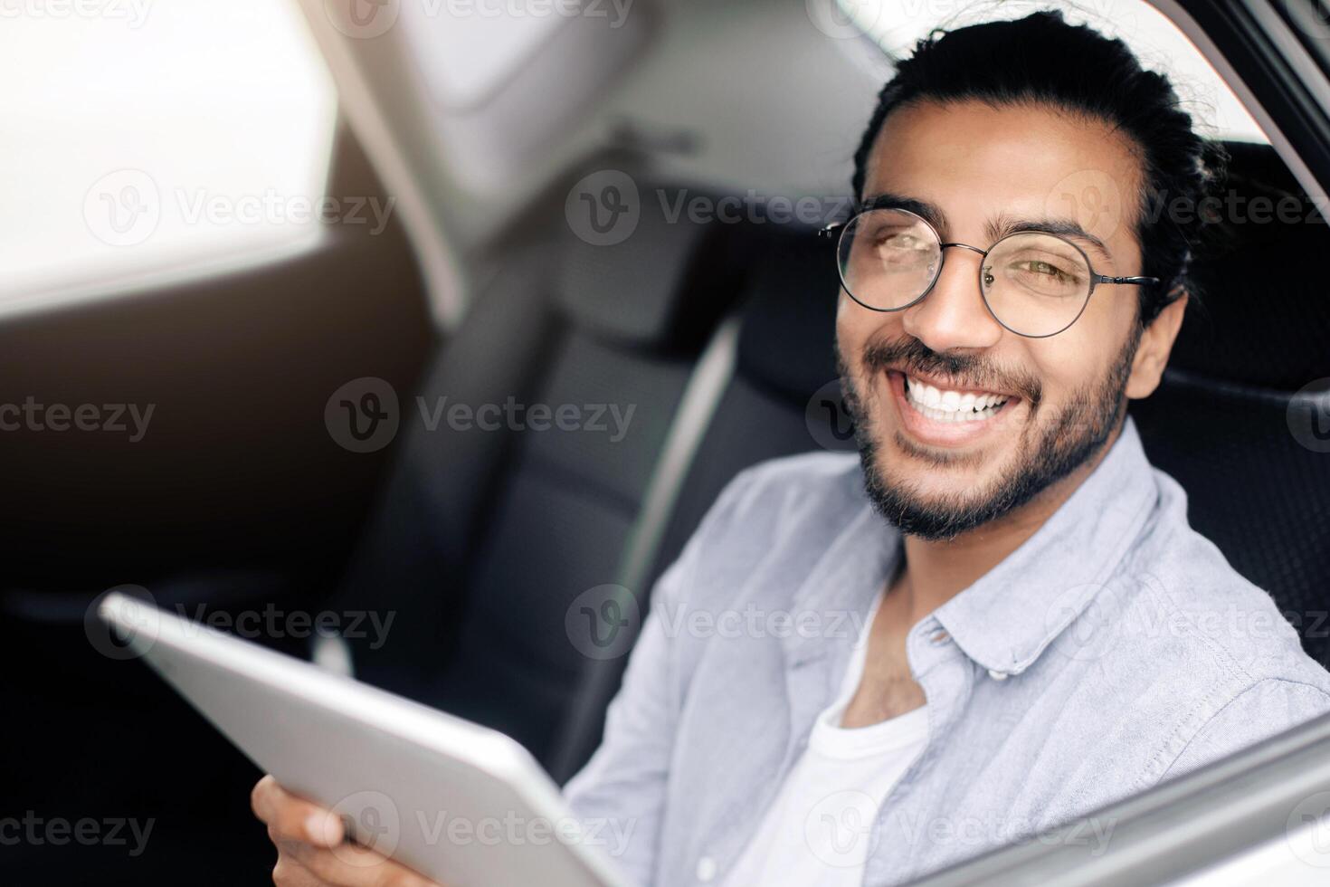Cheerful indian guy using digital tablet while taking cab, copy space. Smiling middle-eastern young man having car trip, sitting on back seat and reading blog on pad. Transportation service concept photo
