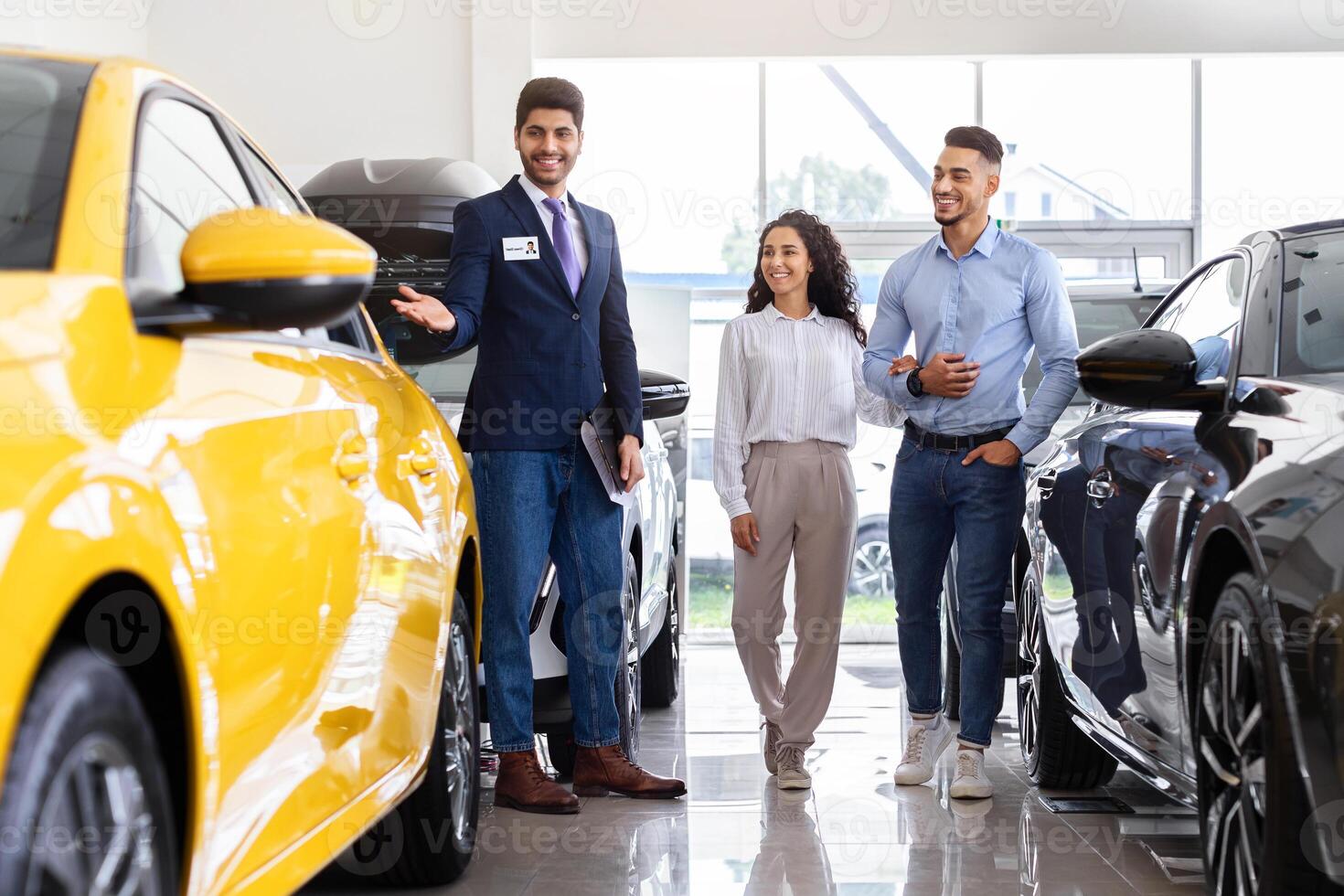 Wealthy middle-eastern young couple having conversation with good-looking sales assistant at auto showroom, looking at nice yellow car and smiling, full length photo, panorama photo