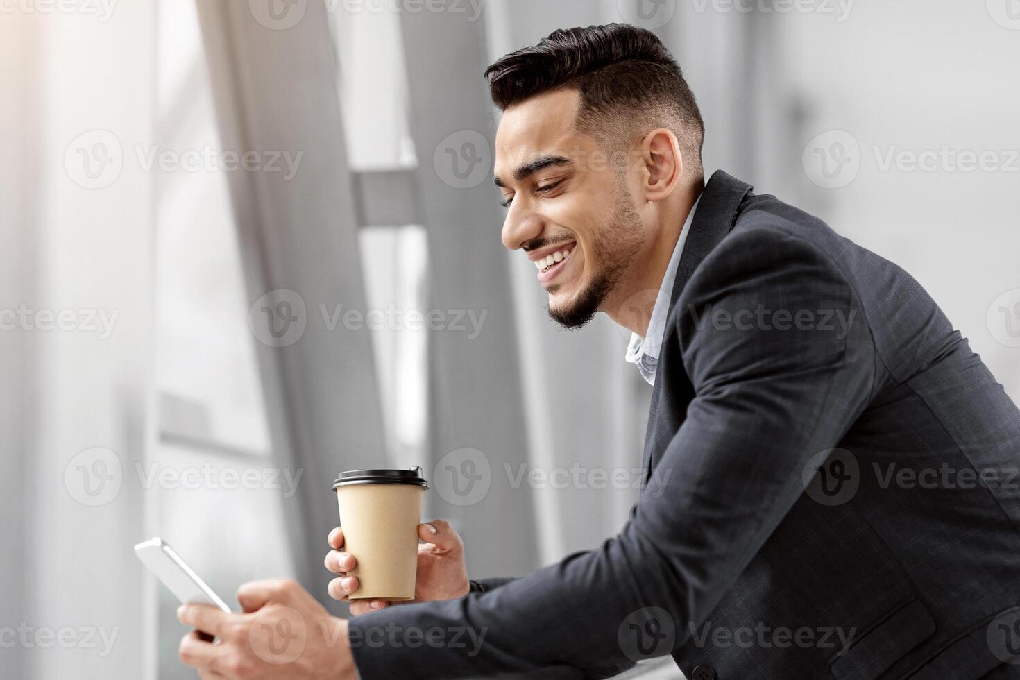 Happy Middle Eastern Businessman Using Smartphone And Having Coffee While Waiting In Airport Terminal, Handsome Arab Man Browsing Internet On Cellphone Or Answering Emails And Enjoying Takeaway Drink photo